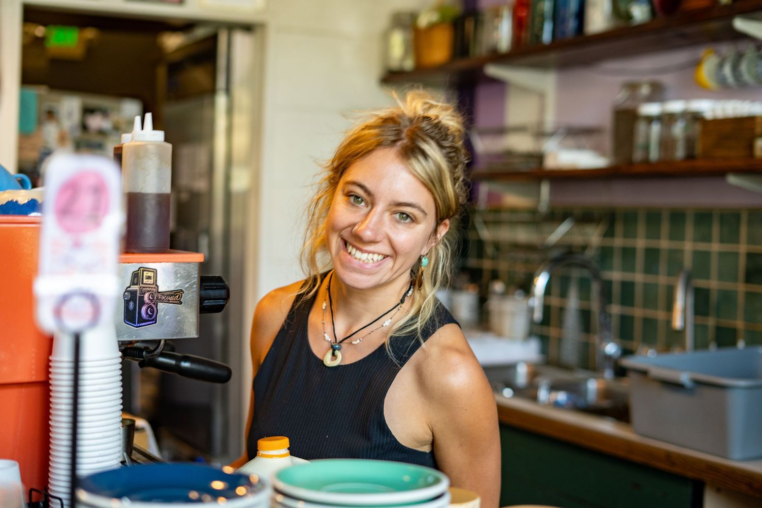 Smiling female barista working behind a coffee shop counter in a casual cafe setting.