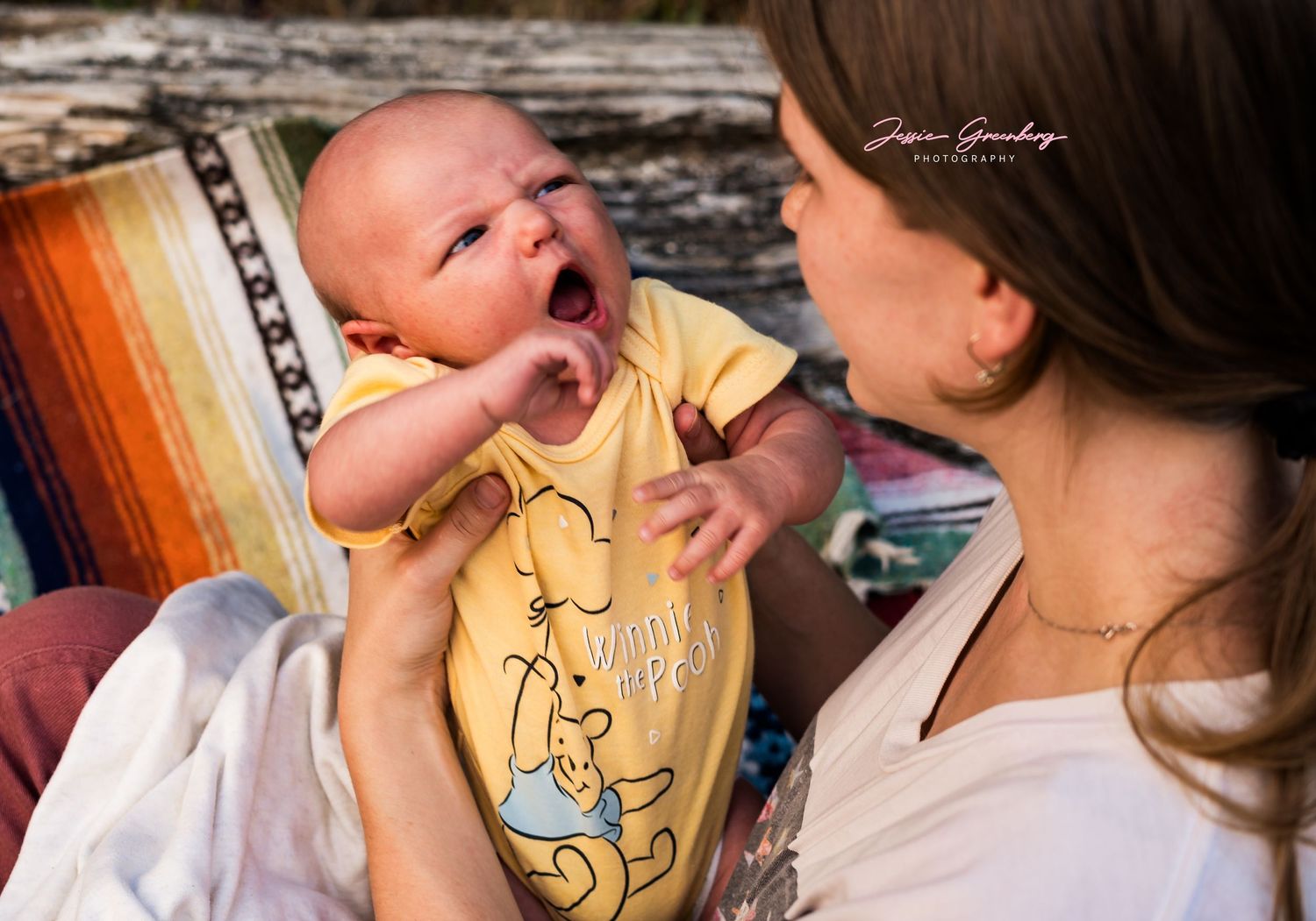 A mother holds her yawning newborn baby dressed in a yellow Winnie the Pooh onesie during an outdoor photo session.