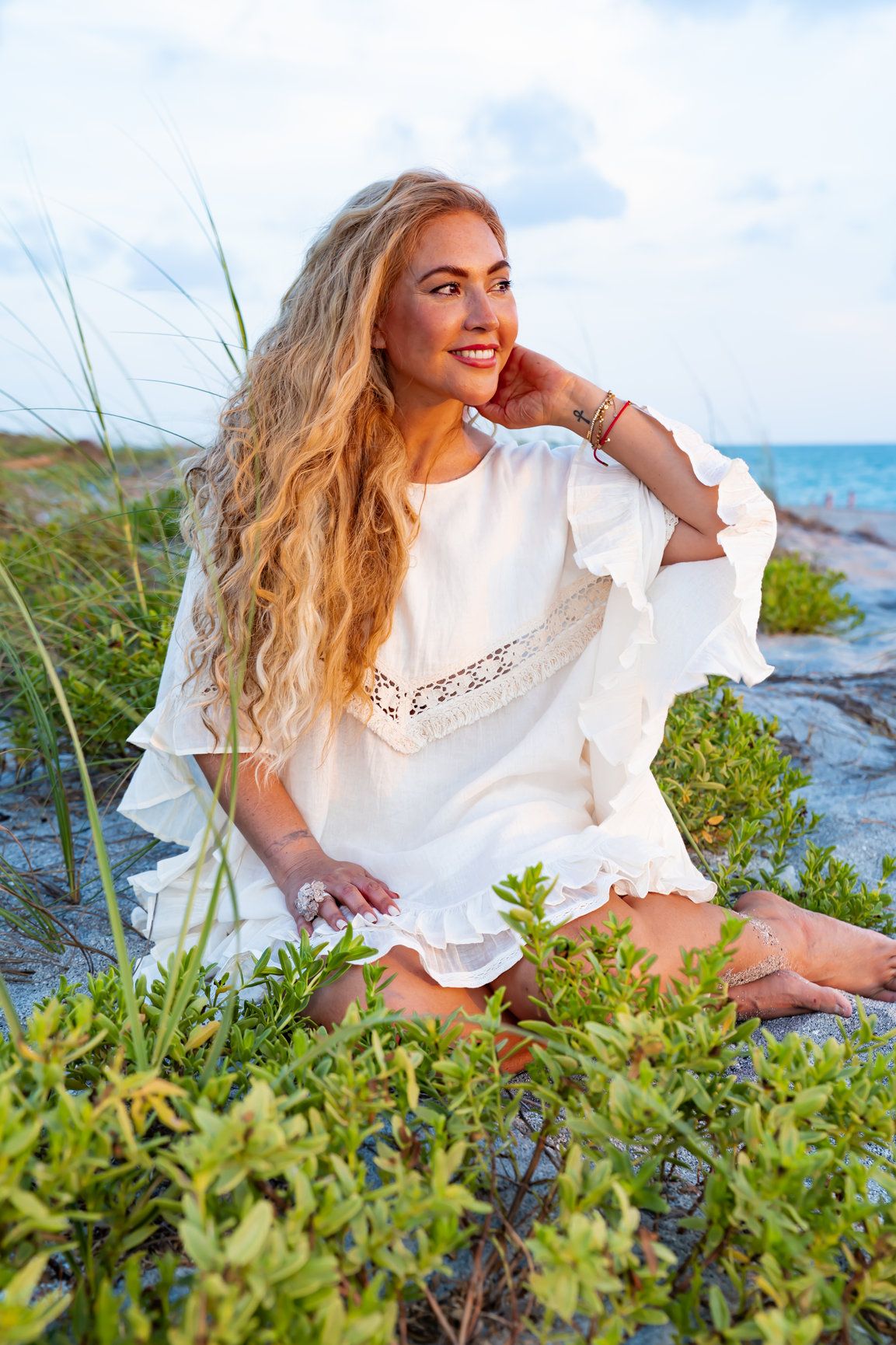 A photo of a woman with blonde flowy hair wearing a white outfit and sitting on greenery on the beach.