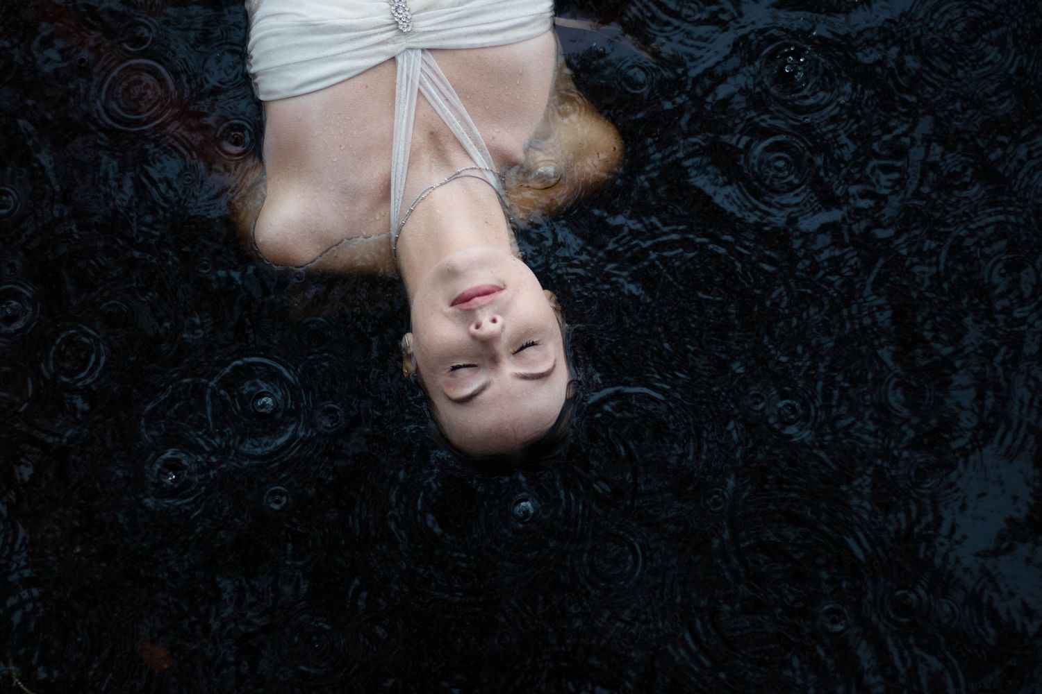 Woman in white dress floating in dark rippling water, viewed from above with serene expression.