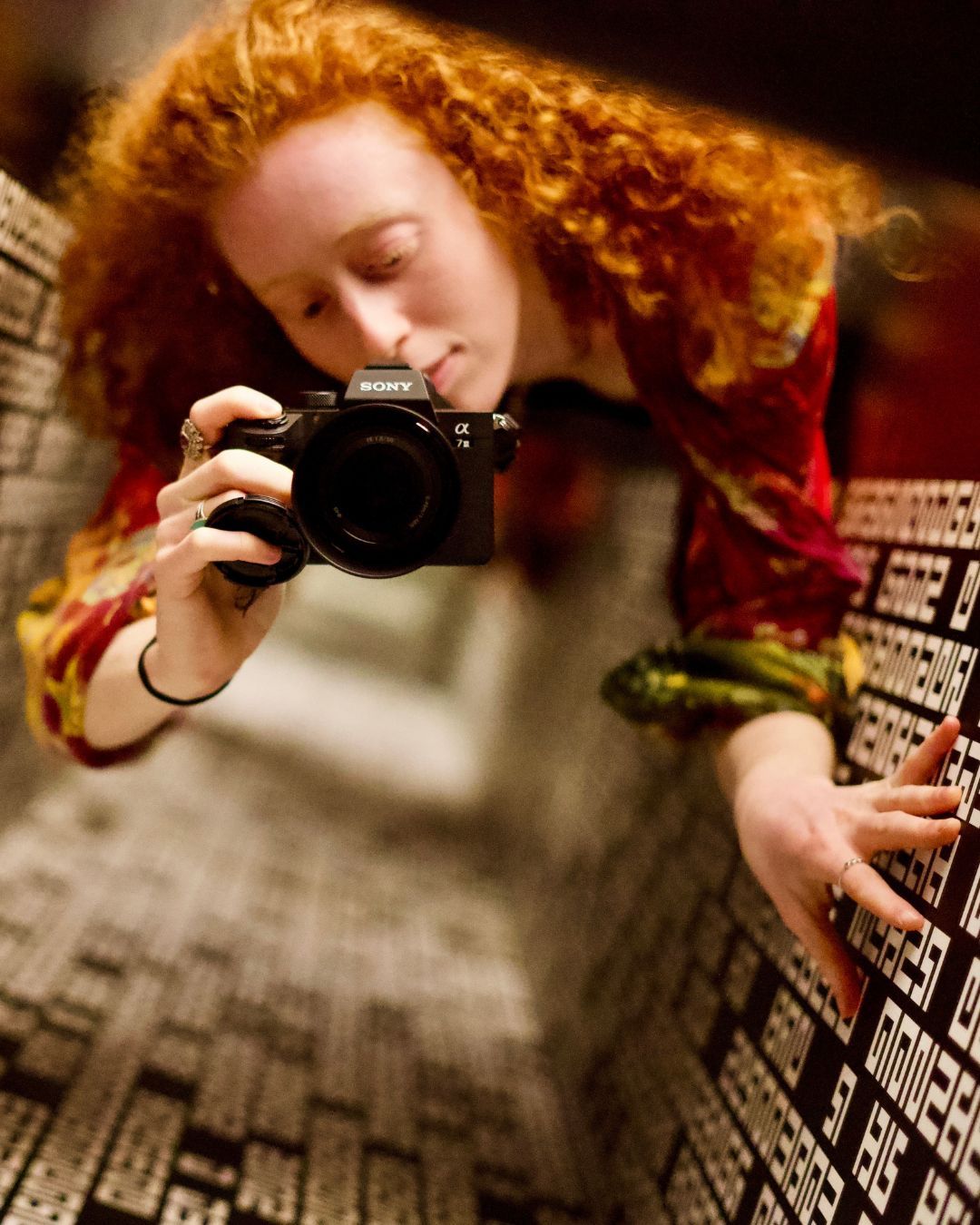 Red-haired woman photographing herself in a mirror with a Sony Alpha camera, wearing a red jacket.