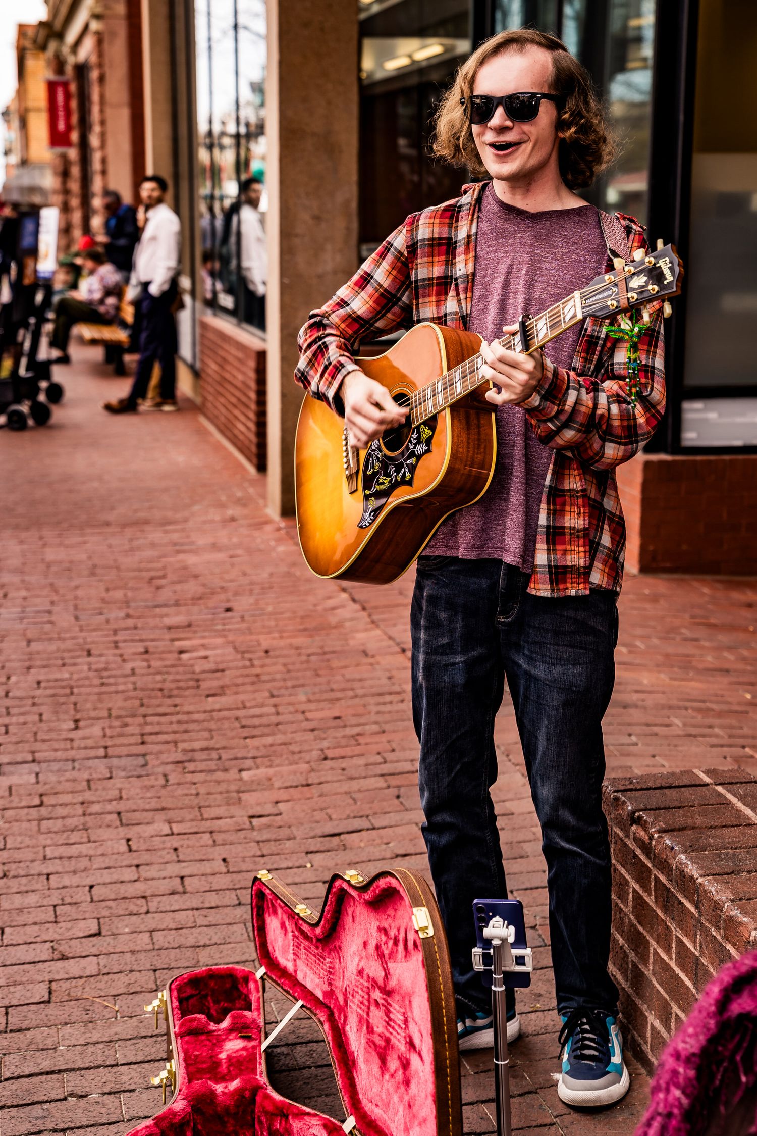 Young male street musician with curly red hair playing acoustic guitar on a brick sidewalk with open guitar case.