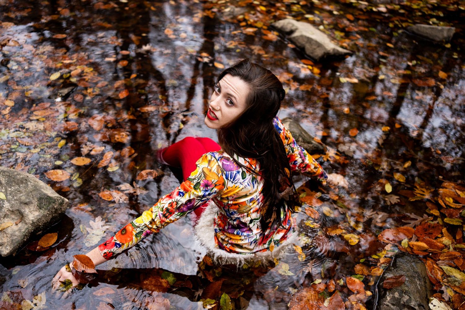 A person in autumn leaves poses near a stream while wearing a colorful floral dress and flowing dark hair.