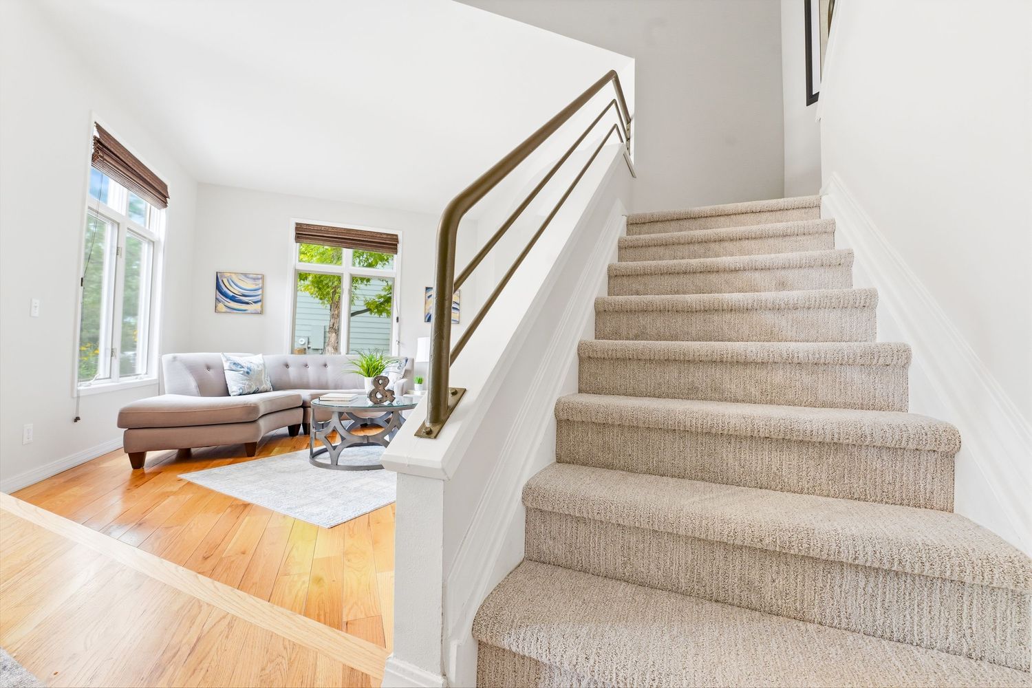Bright entryway with carpeted staircase, white railings, and hardwood floors leading to a sunlit living room with sofa.