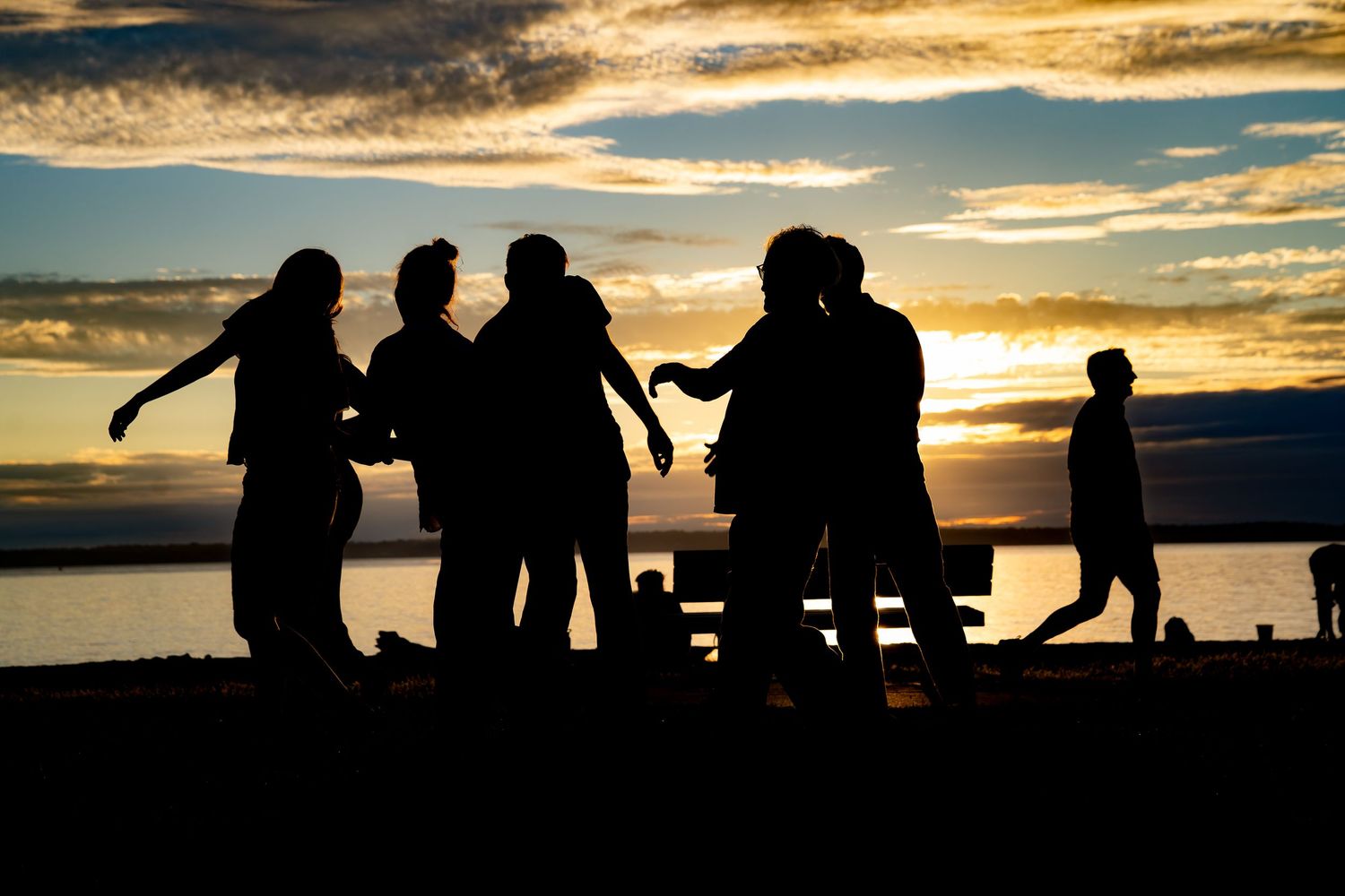 Silhouettes of a group of friends gathered around a picnic table at sunset by the water.