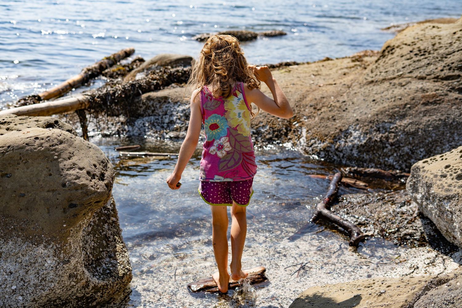 A young girl in a colorful dress explores rocky shoreline tide pools, splashing in shallow water near the ocean.