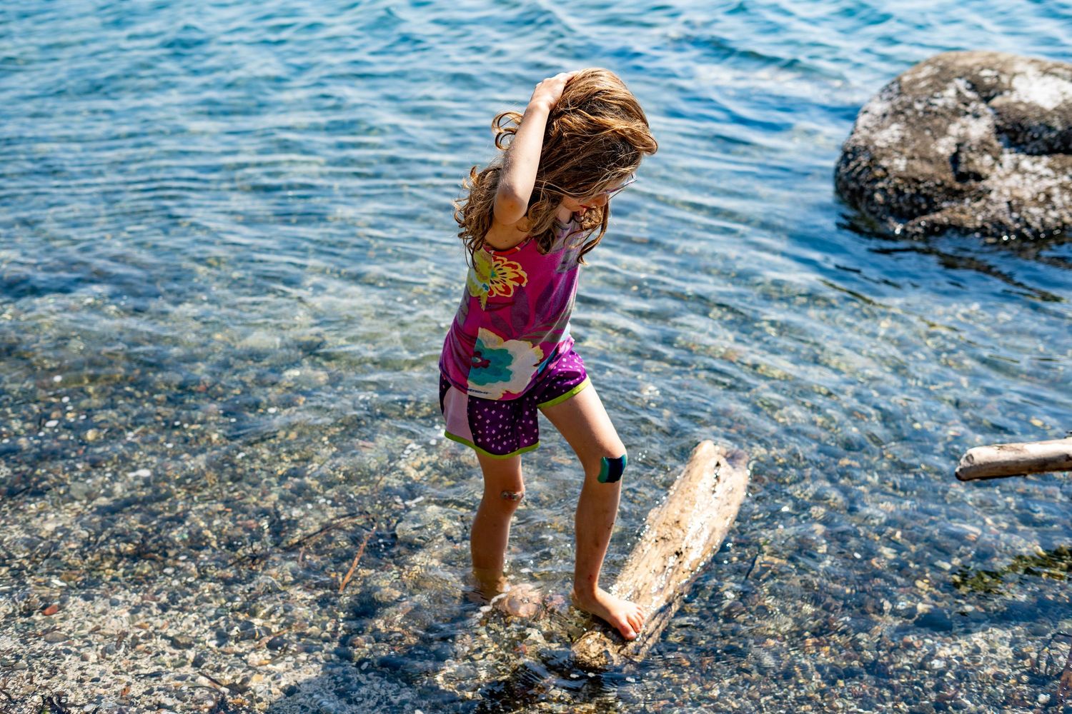 Young girl in colorful swimsuit stepping on driftwood along a rocky shoreline with clear blue water.