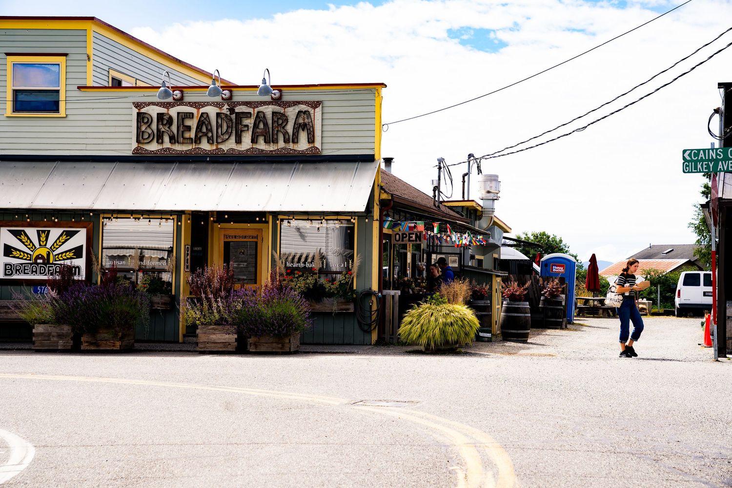 Person walking past Breadfarm bakery storefront in a small town with colorful signage and outdoor planters.