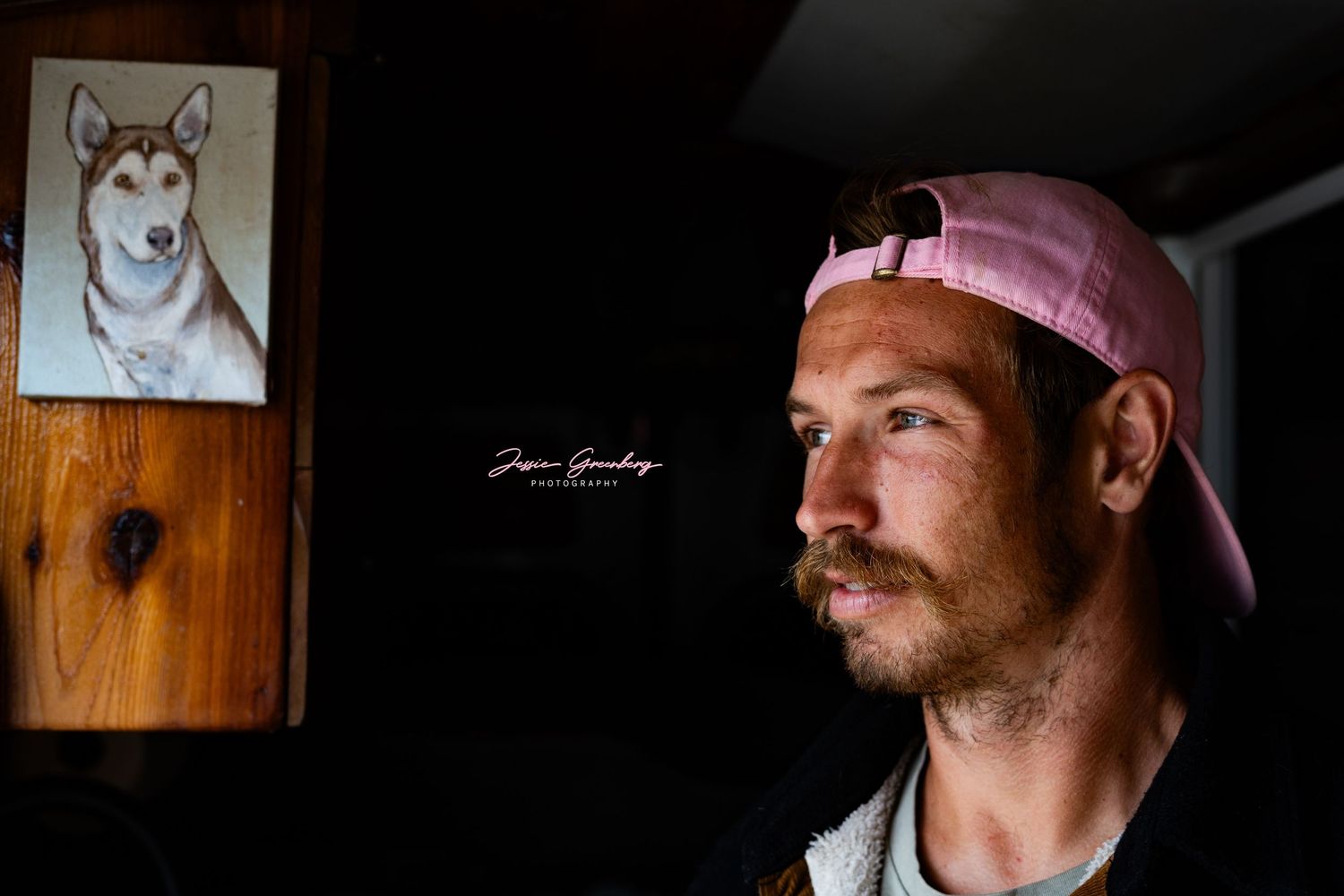 Man in pink cap poses indoors beside a framed husky dog portrait painting on a wooden wall.