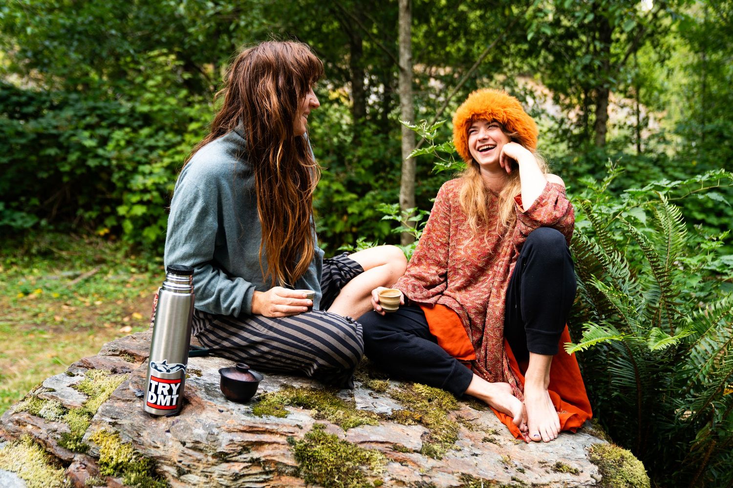 Two women laughing together on a mossy rock outdoors, surrounded by lush green forest trees.
