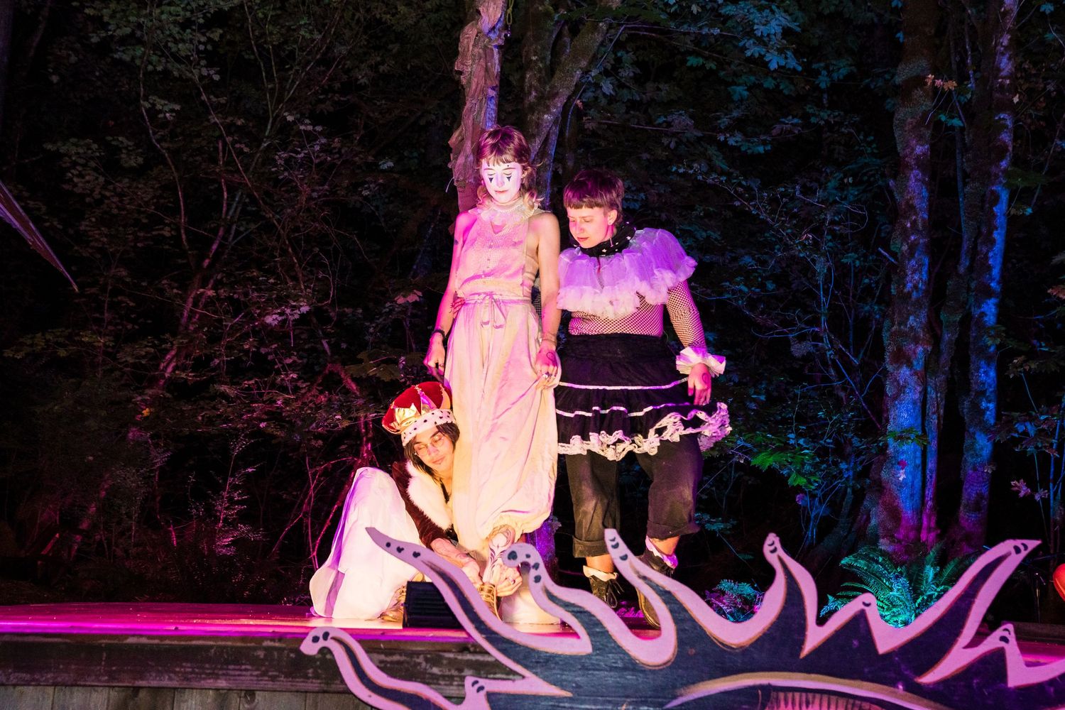 Three cosplayers pose on a colorful illuminated stage outdoors at night, dressed in vibrant fantasy costumes.