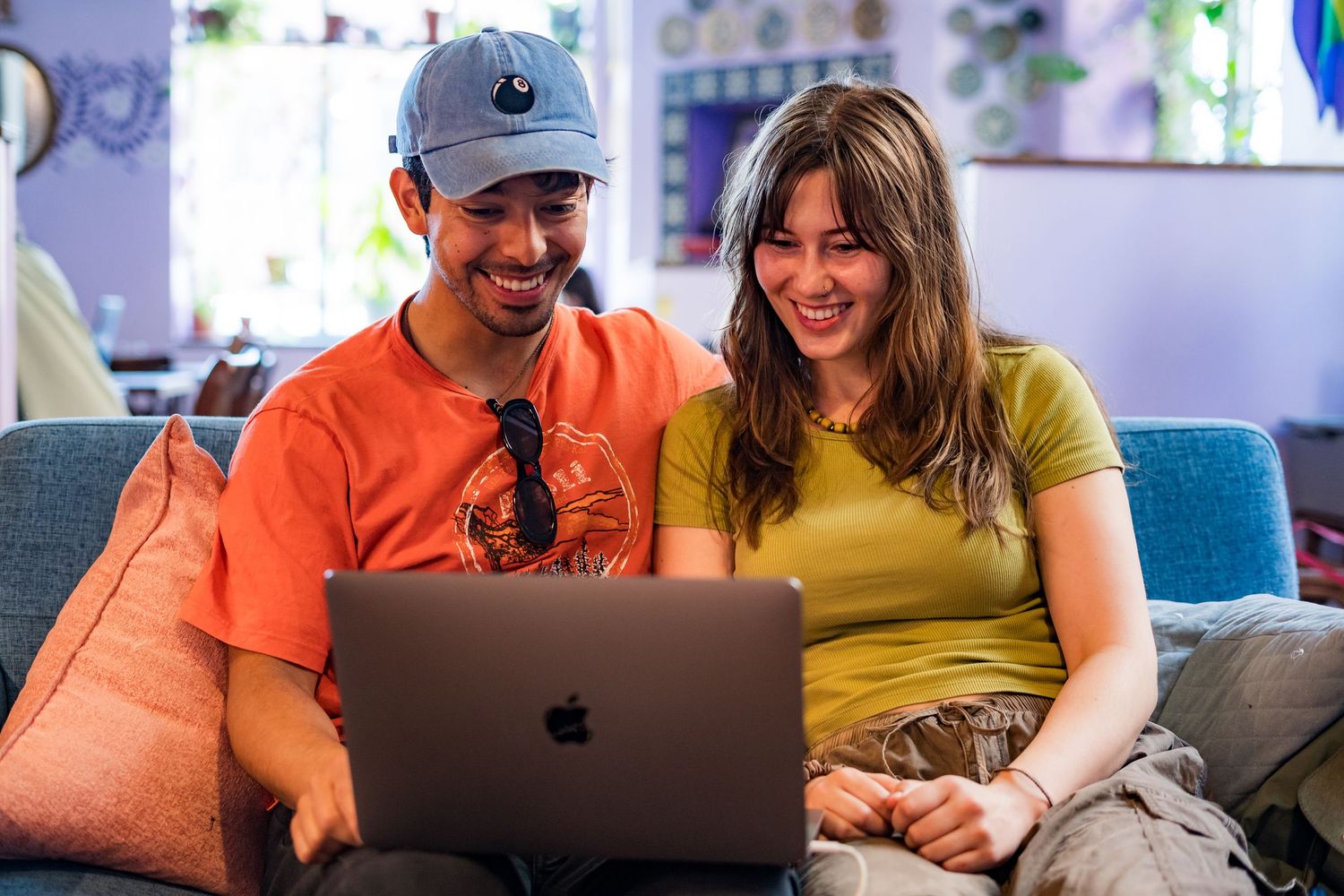 Two friends smiling and laughing together while browsing on a MacBook laptop seated on a cozy blue sofa.