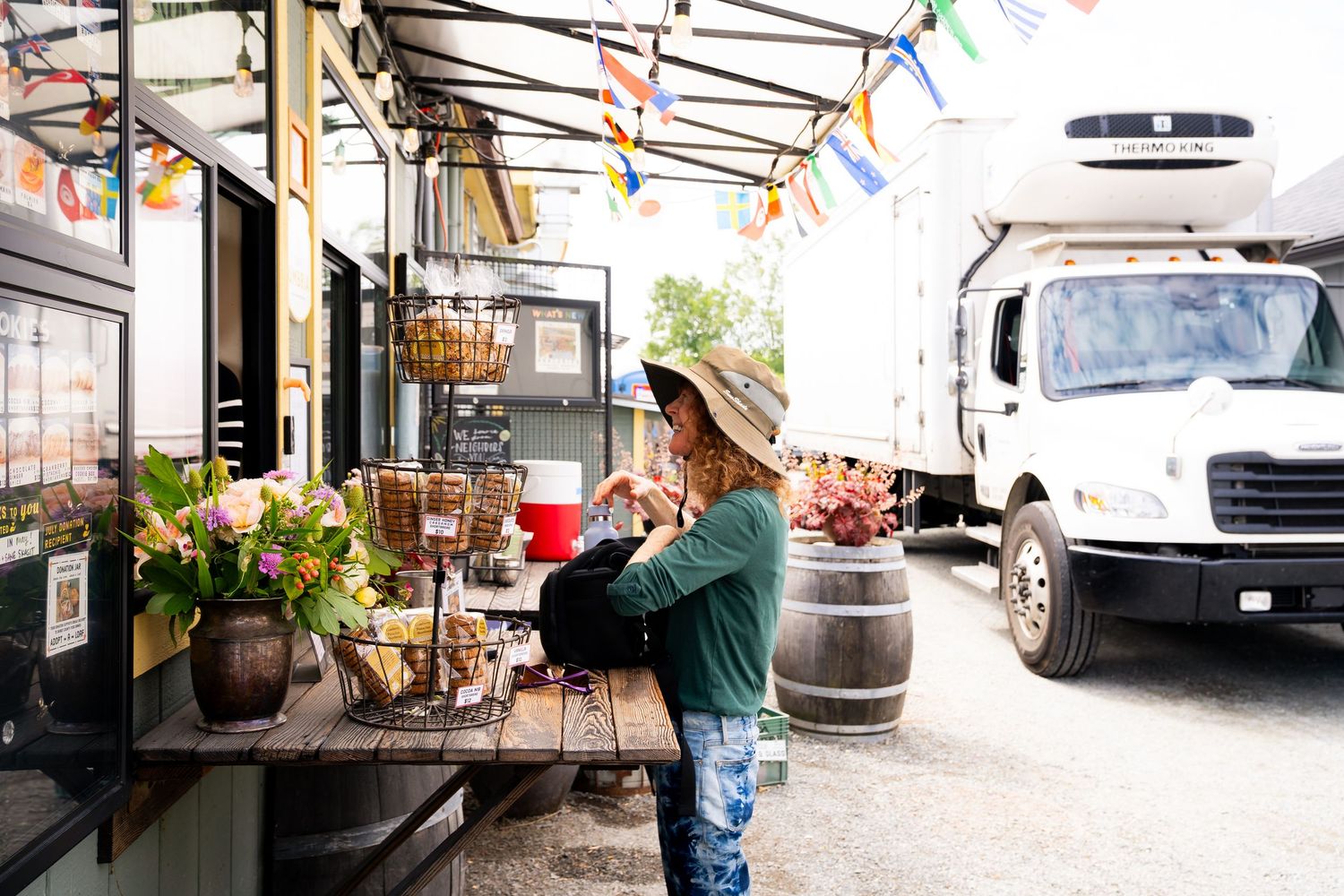 Woman in hat browsing colorful outdoor food truck market with floral decor and delivery truck in background.