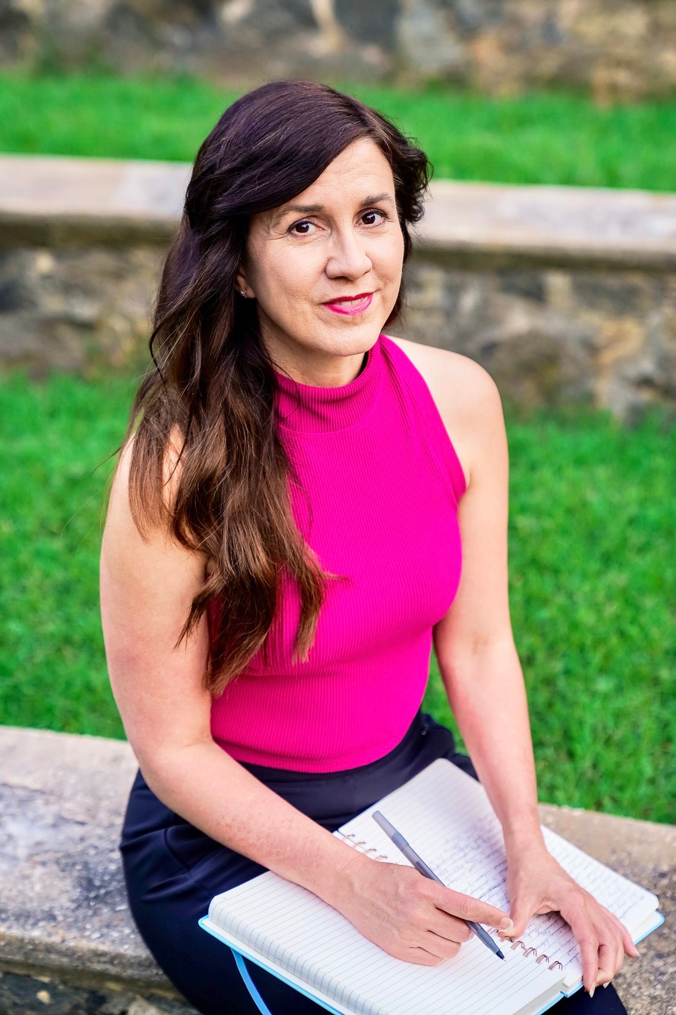 A person in a bright pink sleeveless top writes in a notebook while sitting outdoors on stone steps with green grass behind.