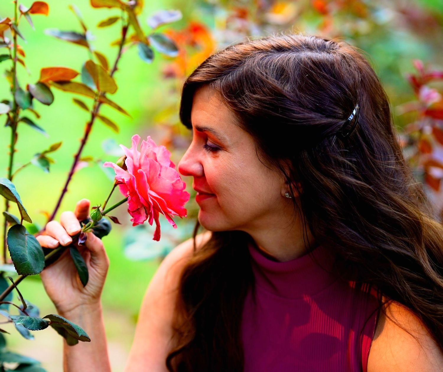 A person in a burgundy sweater smells pink flowers in a garden setting with blurred green foliage in the background.