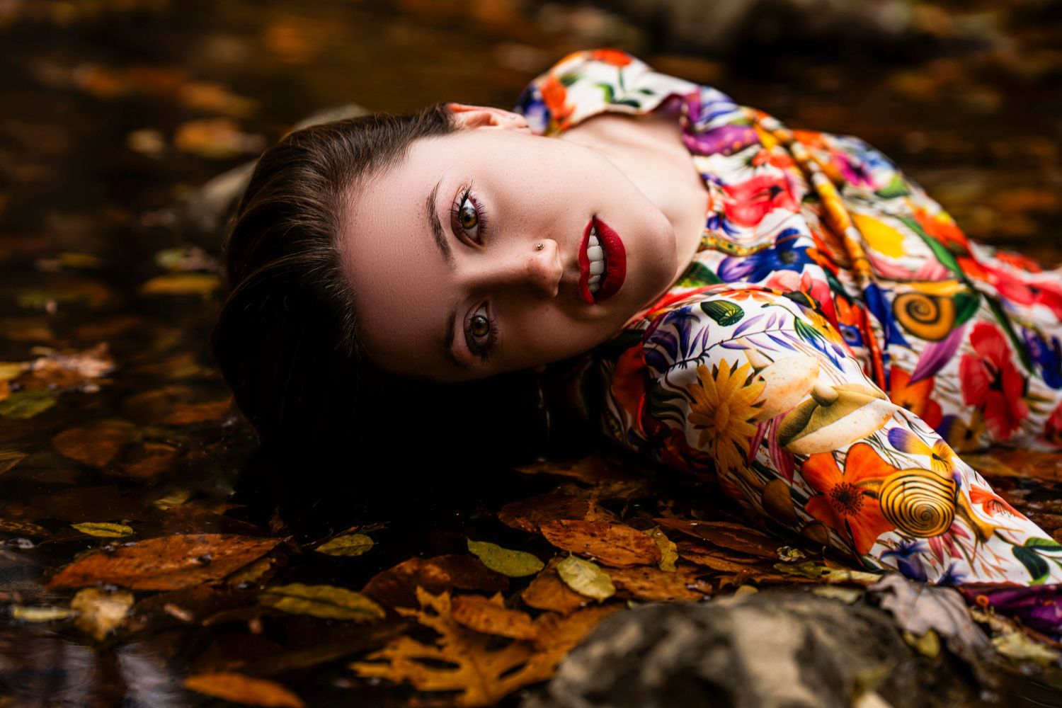 A figure in a colorful floral dress lies gracefully among autumn leaves on the ground.