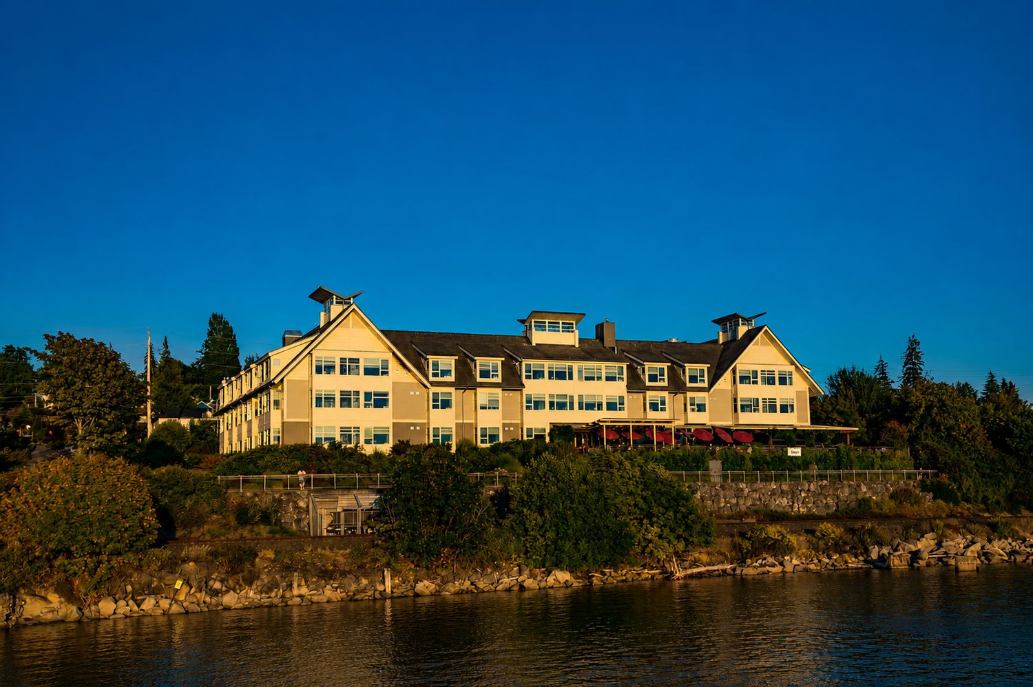 Large yellow waterfront hotel surrounded by trees reflecting golden hour sunlight on calm blue waters.
