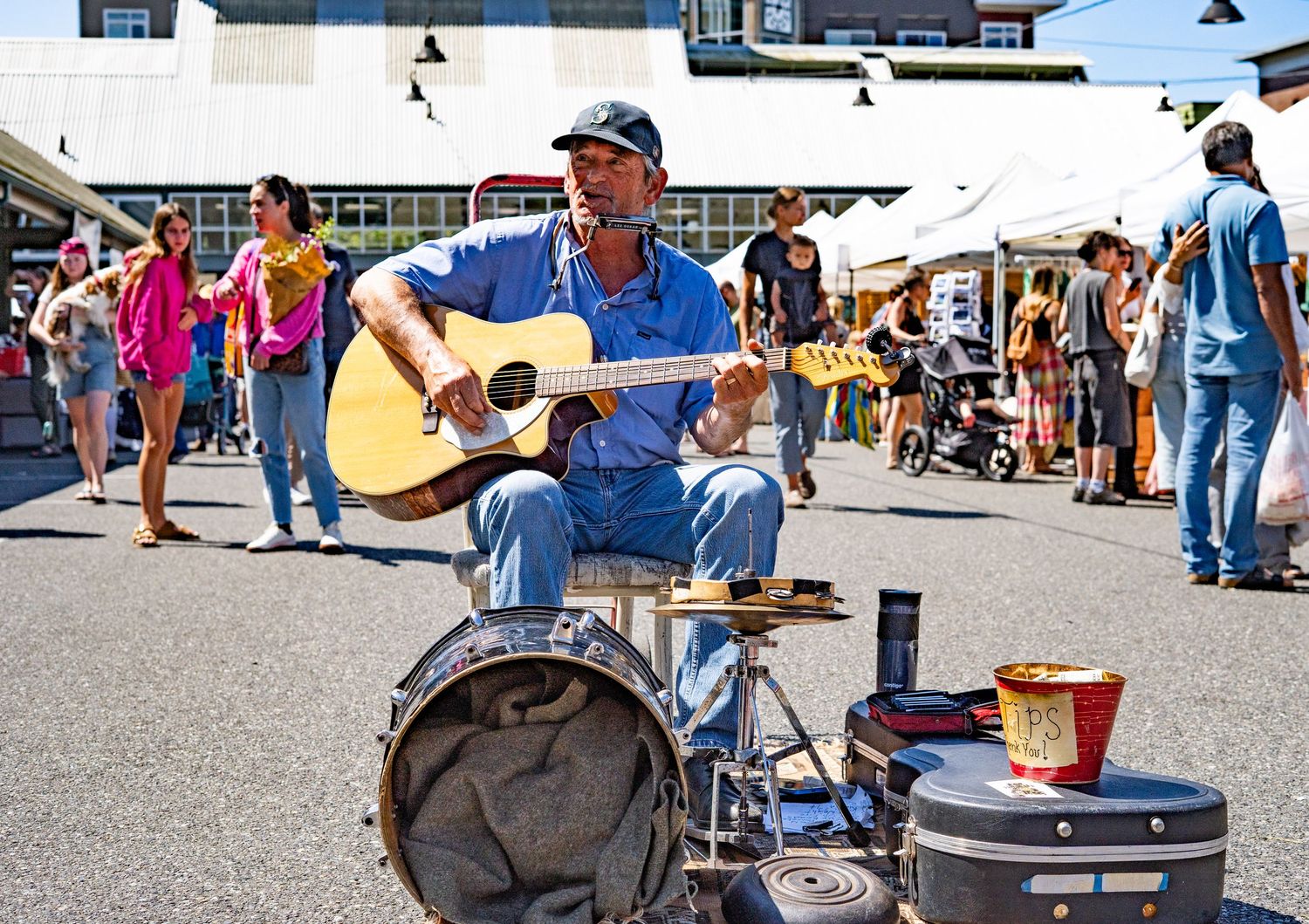 Street musician playing acoustic guitar while seated at a drum kit at an outdoor market on a sunny day.