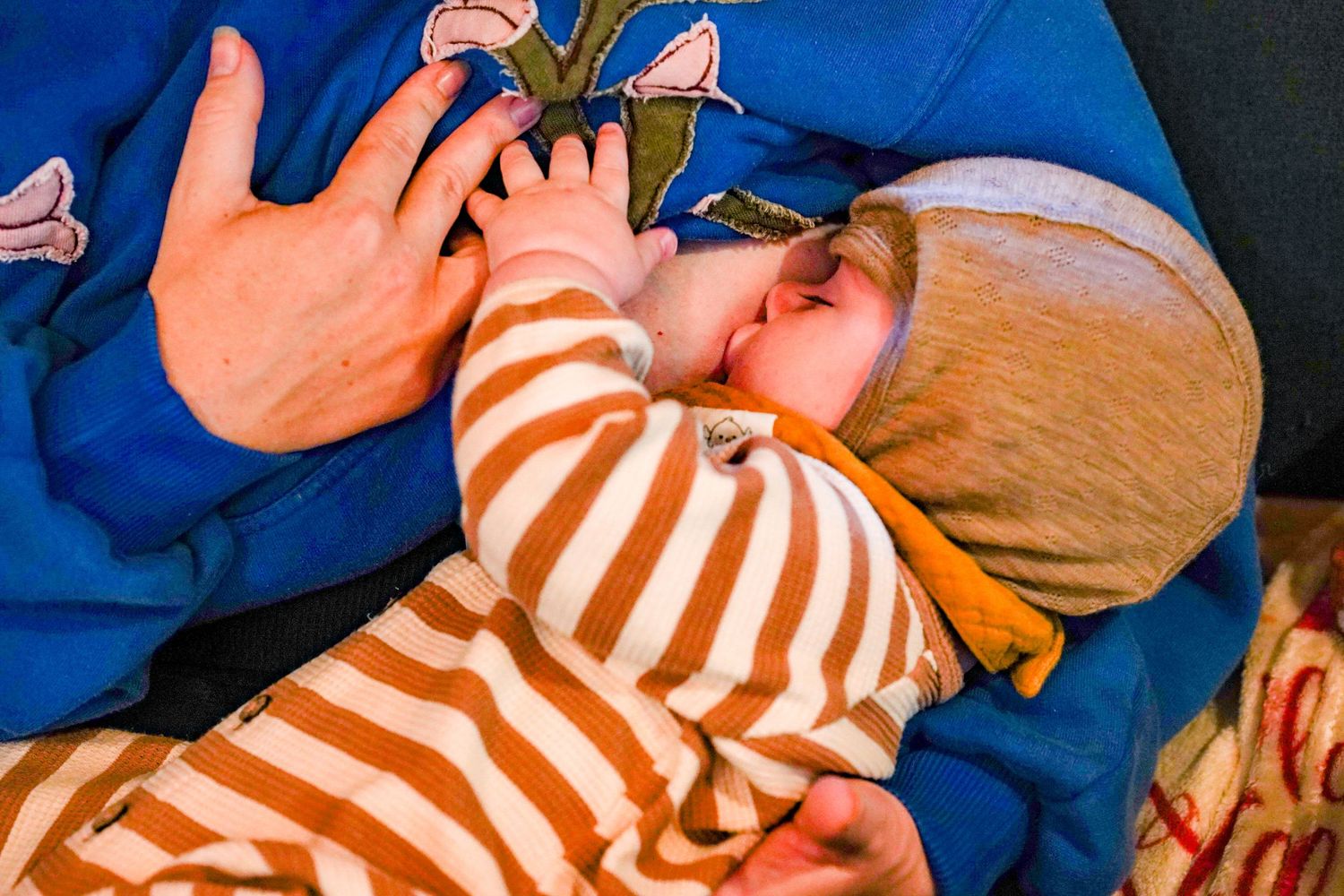 A newborn baby in orange striped pajamas breastfeeding while being cradled by a parent in a blue shirt.