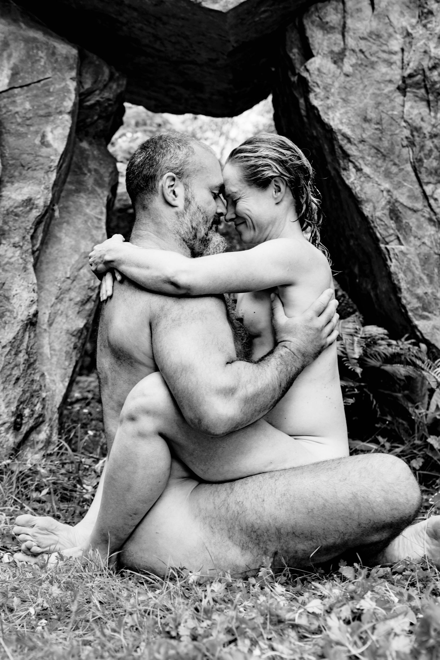 Black and white photo of a nude couple embracing intimately outdoors near rocky formation.
