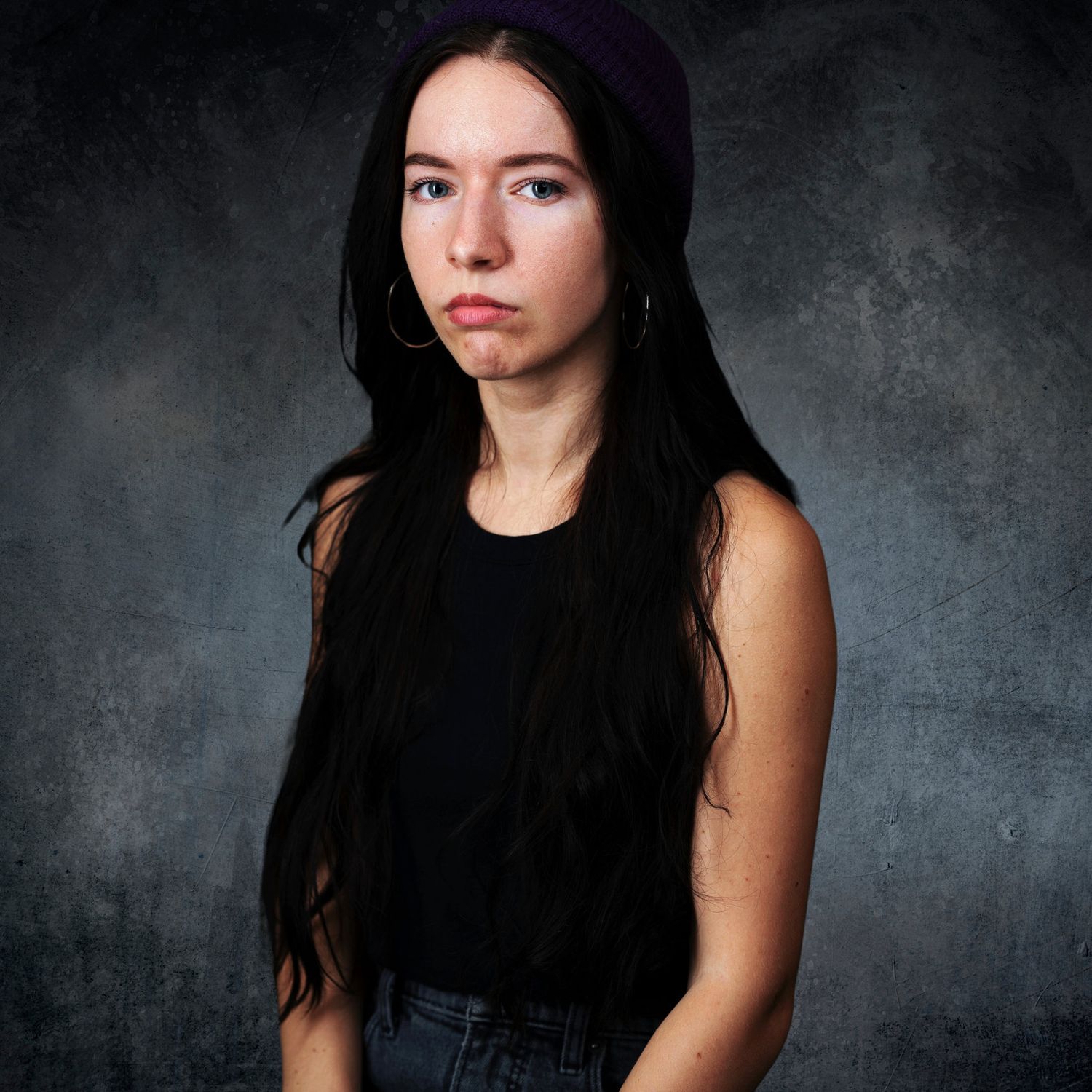 A photo of a girl with long black hair looking at the camera not smiling, this photo was taken in a studio with a blue backdrop.