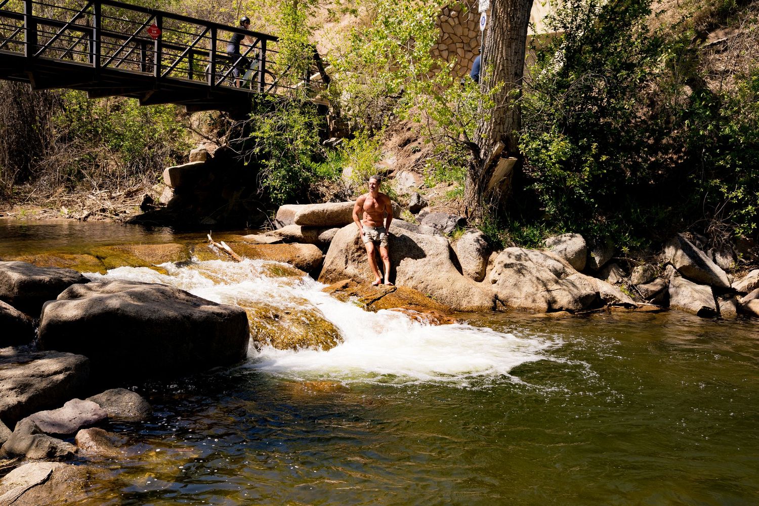 Person sitting on rocks beside a flowing stream with a wooden bridge and lush trees in a natural outdoor setting.