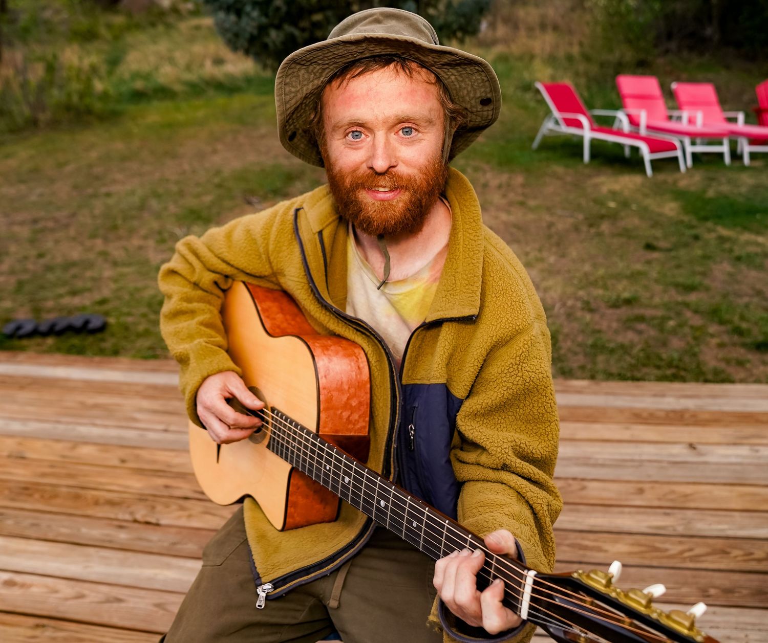 Smiling bearded man in olive fleece and bucket hat playing acoustic guitar on wooden deck outdoors.