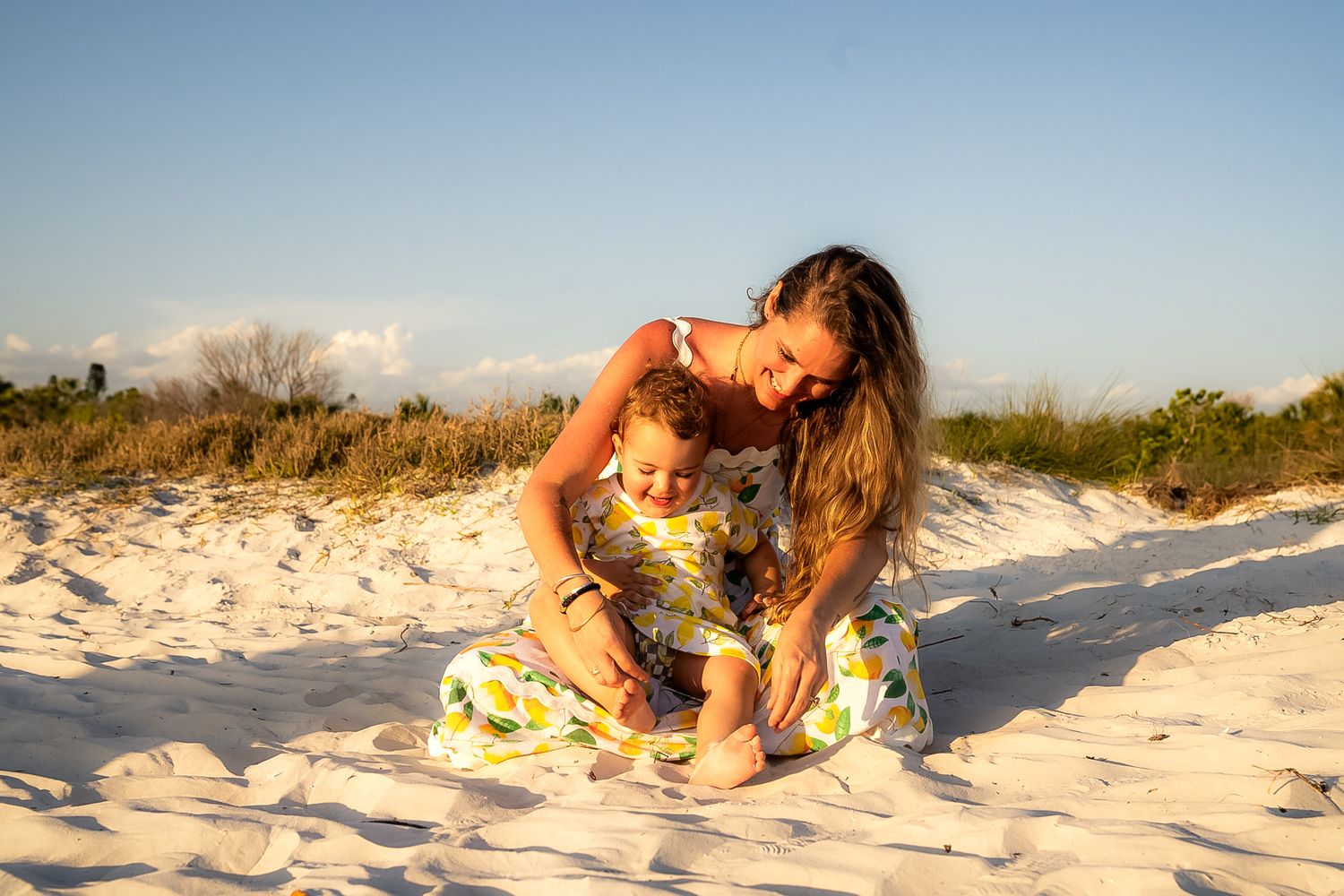 A photo of a woman holding a baby girl sitting on the sand, they were smiling at each other.