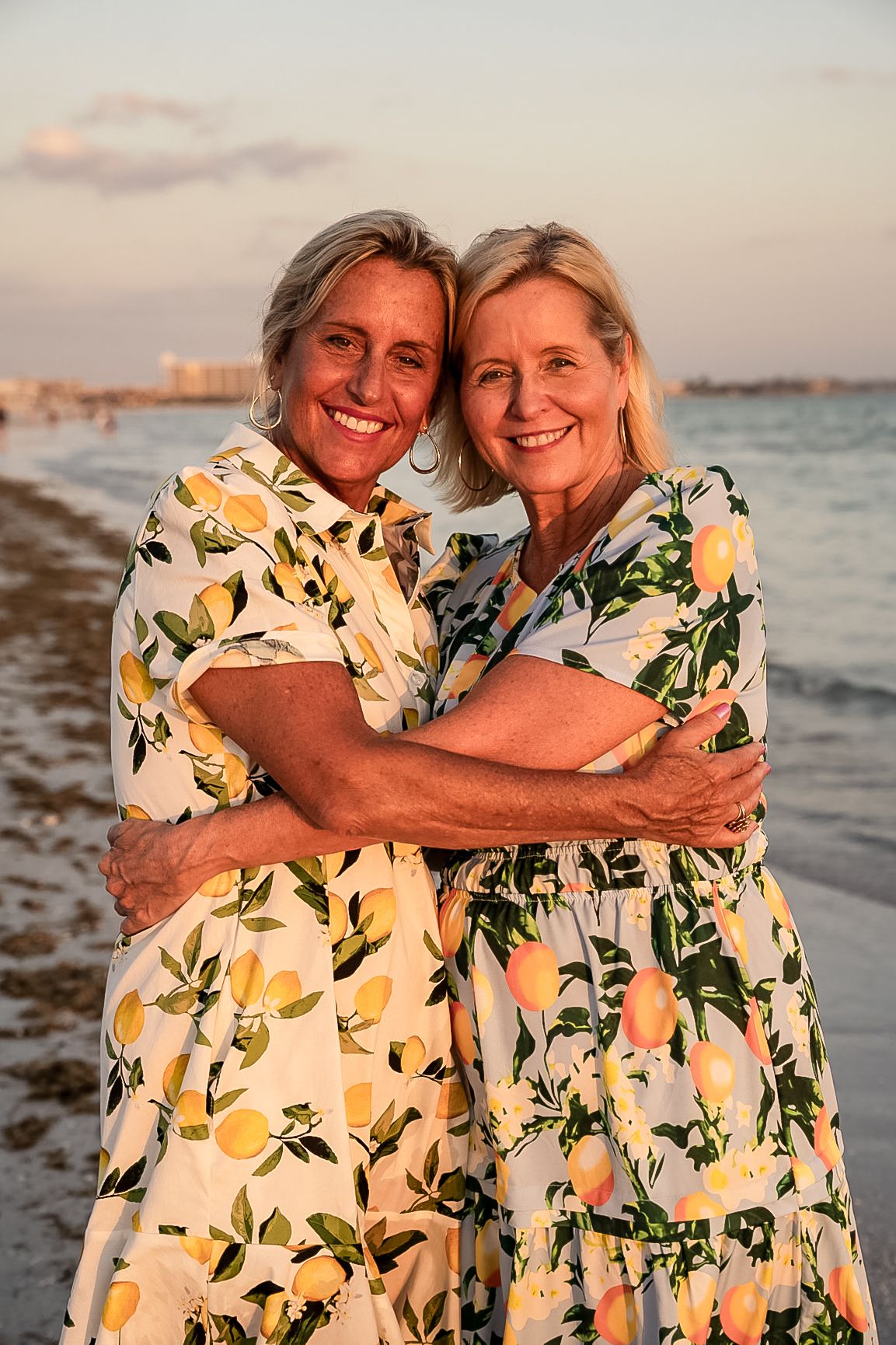 A photo of the two sisters hugging on the beach with patterned dresses and water behind them.