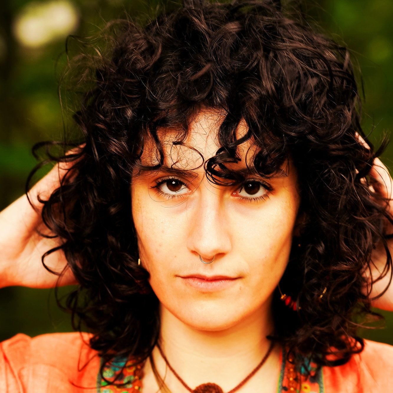 A photo of a woman looking intensely at the camera, she is wearing an apricot top and her hands are on her head.