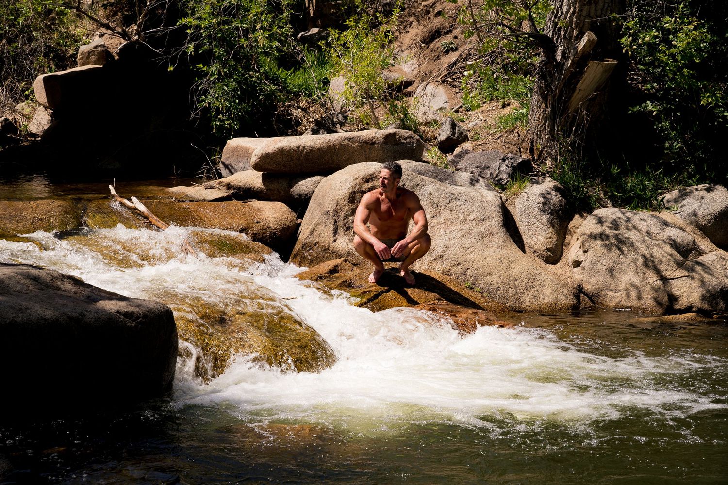 Shirtless man sitting on rocks beside a rushing mountain river waterfall in a scenic outdoor setting.
