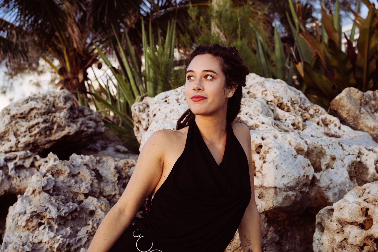 A woman in a black dress poses dramatically against rocky cliffs and palm trees at sunset.