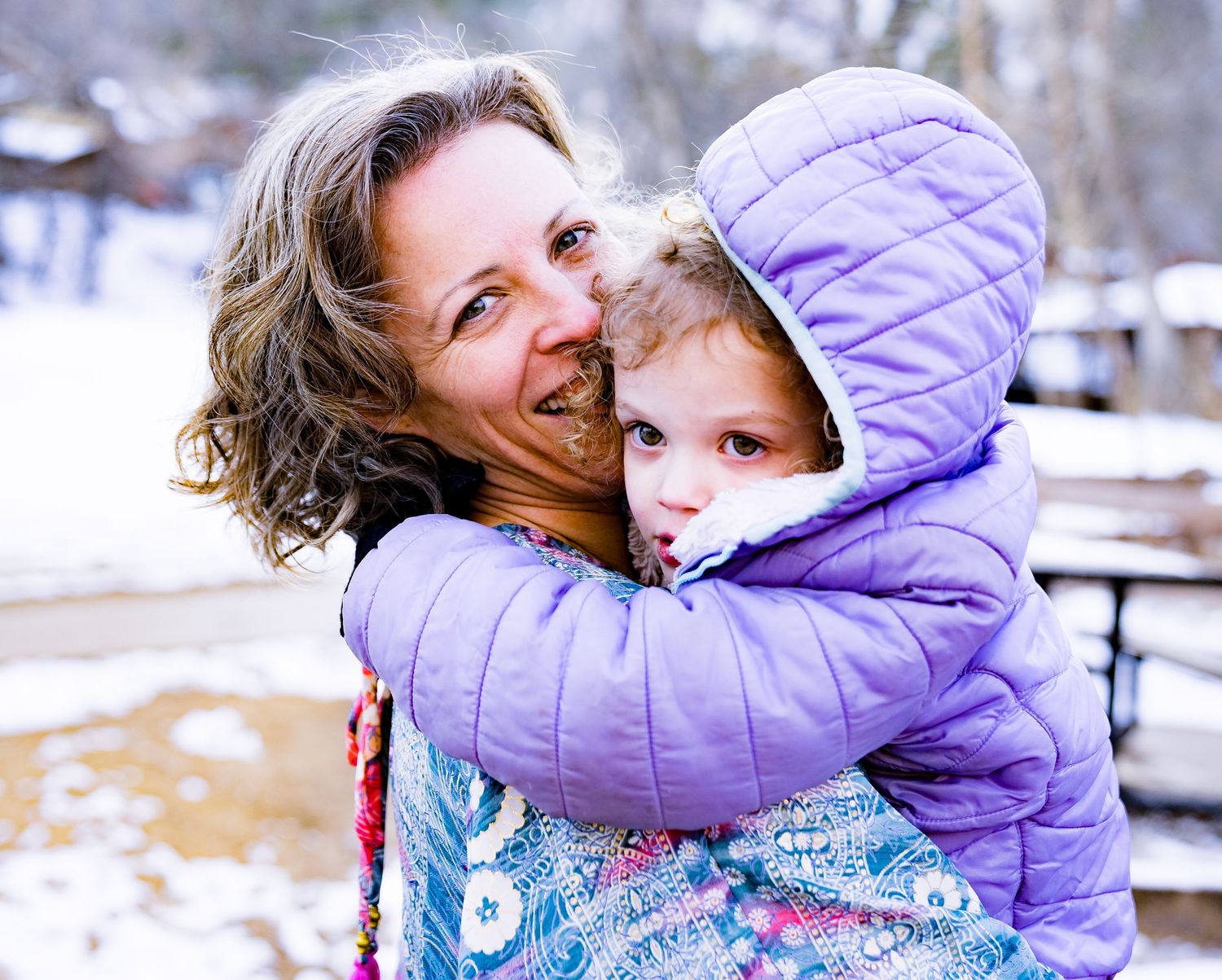 Two people share a warm embrace outdoors in winter with snow visible in the background.
