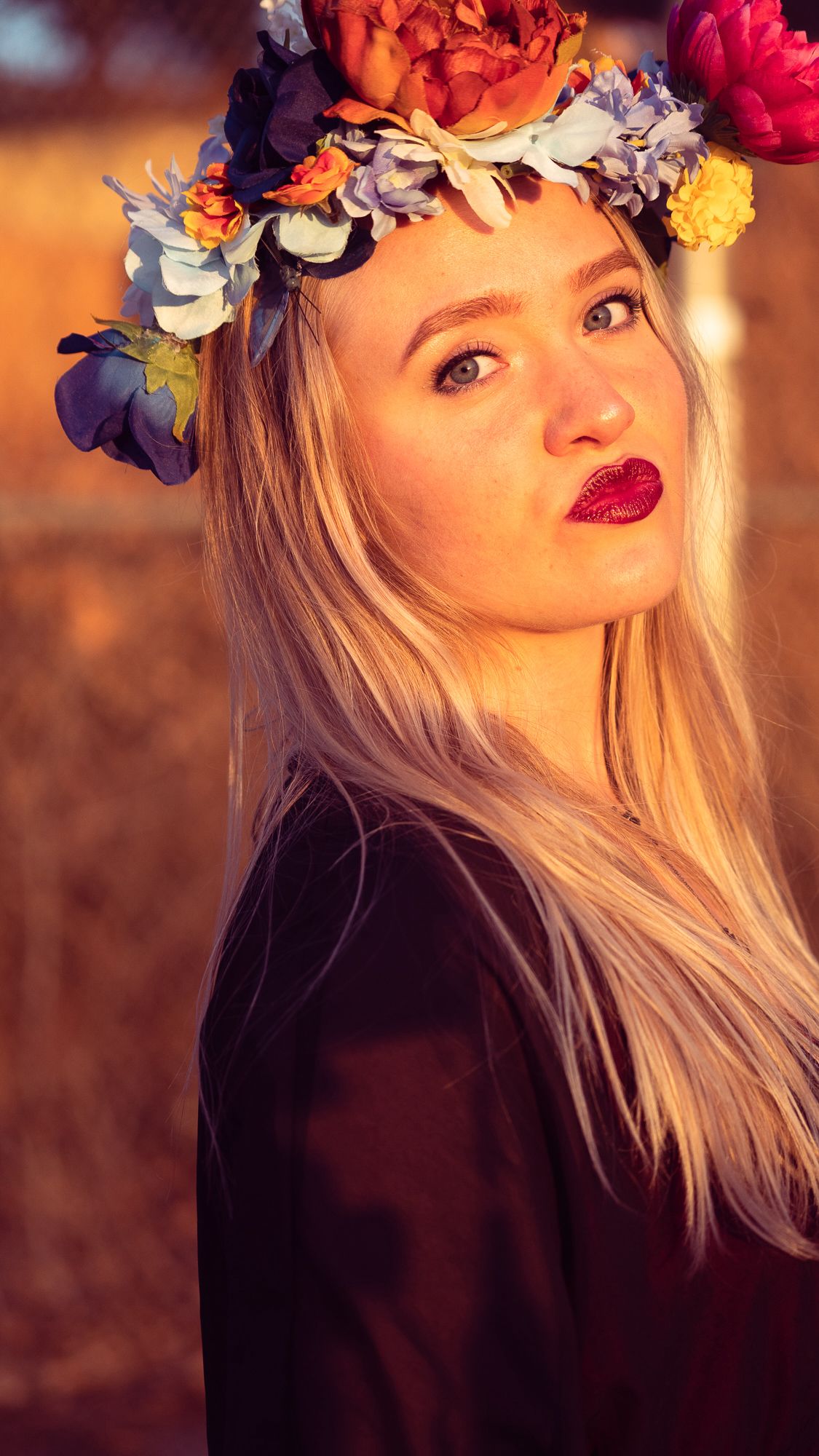 Person wearing vibrant flower crown with red and purple blooms poses against warm sunset lighting in artistic portrait.