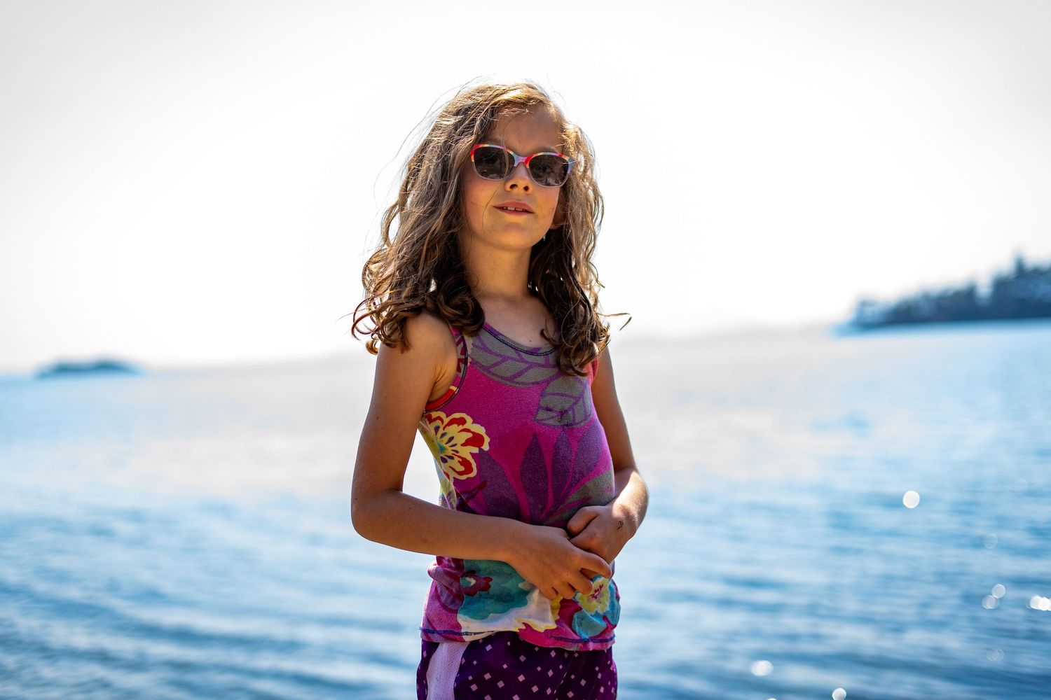 A young girl with curly hair wearing sunglasses and a colorful dress stands near a blue ocean on a sunny day.