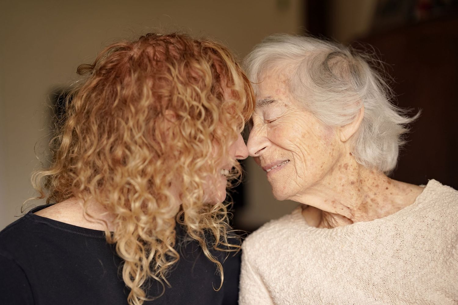 An elderly woman and younger curly red-haired woman share a tender forehead-to-forehead moment indoors.