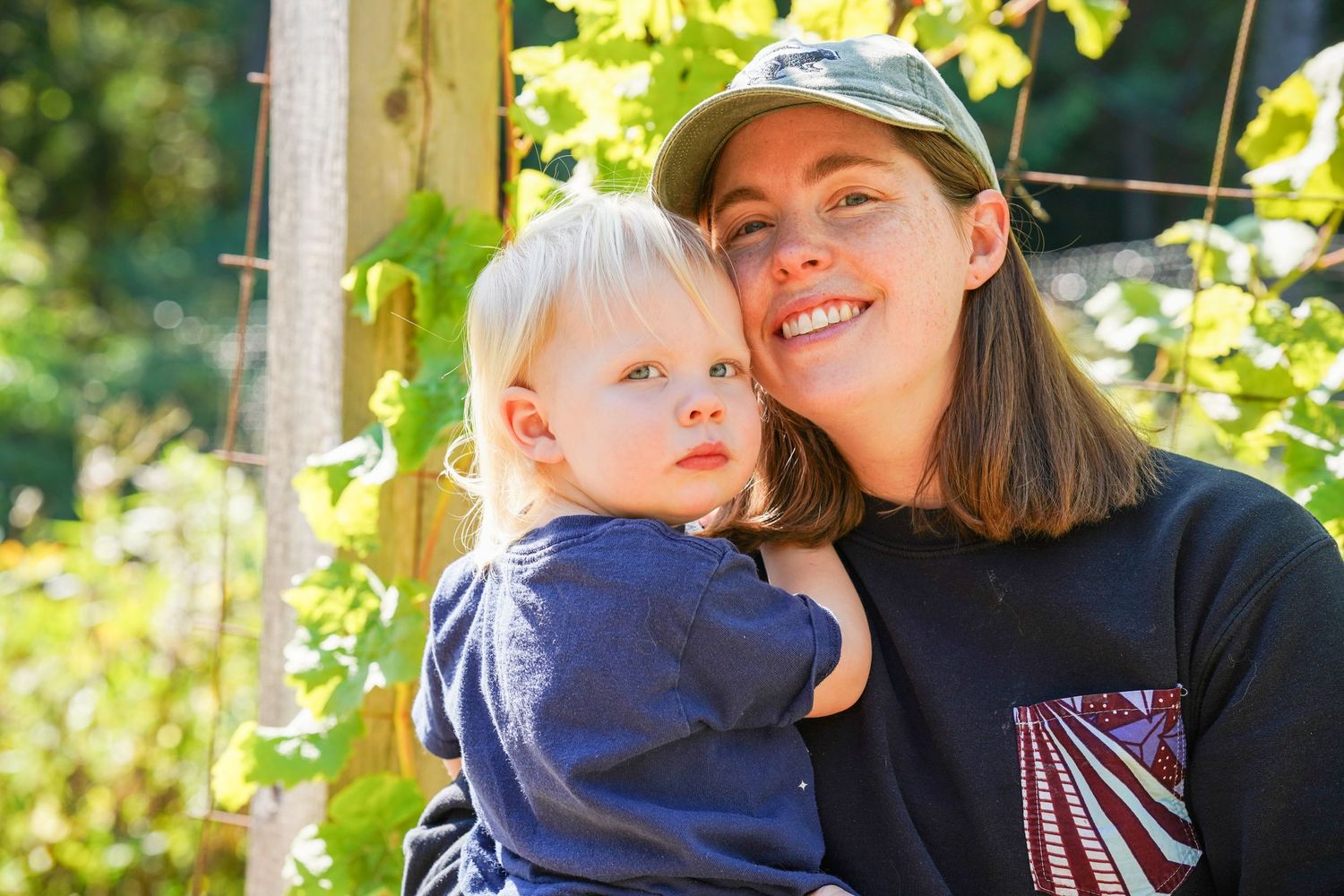 A smiling woman in a green cap holds a young blonde toddler outdoors among green vines in natural sunlight.