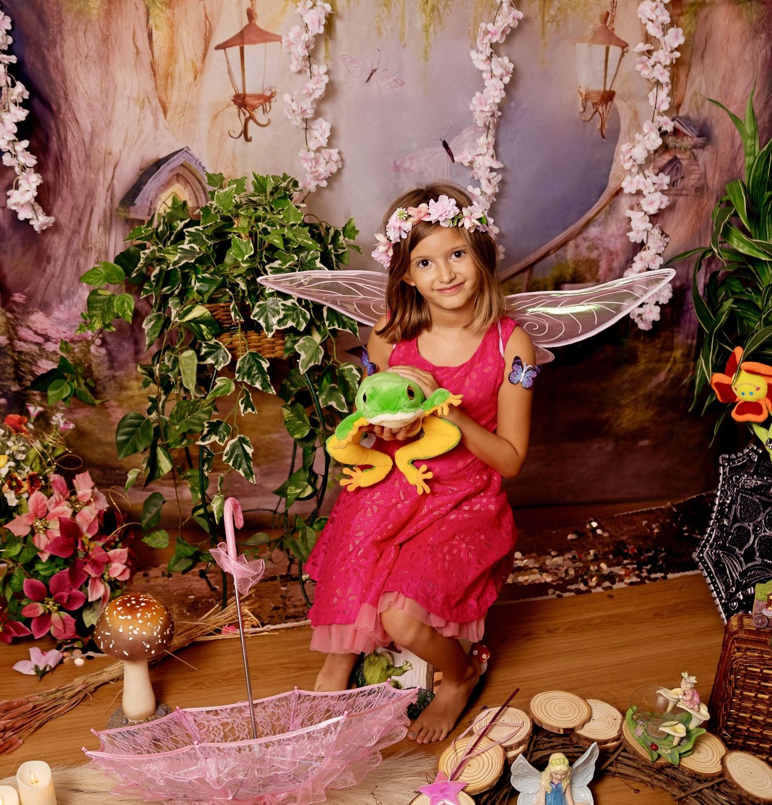 Young girl in pink fairy costume with wings holds a toy frog in an enchanted forest themed photo setting.