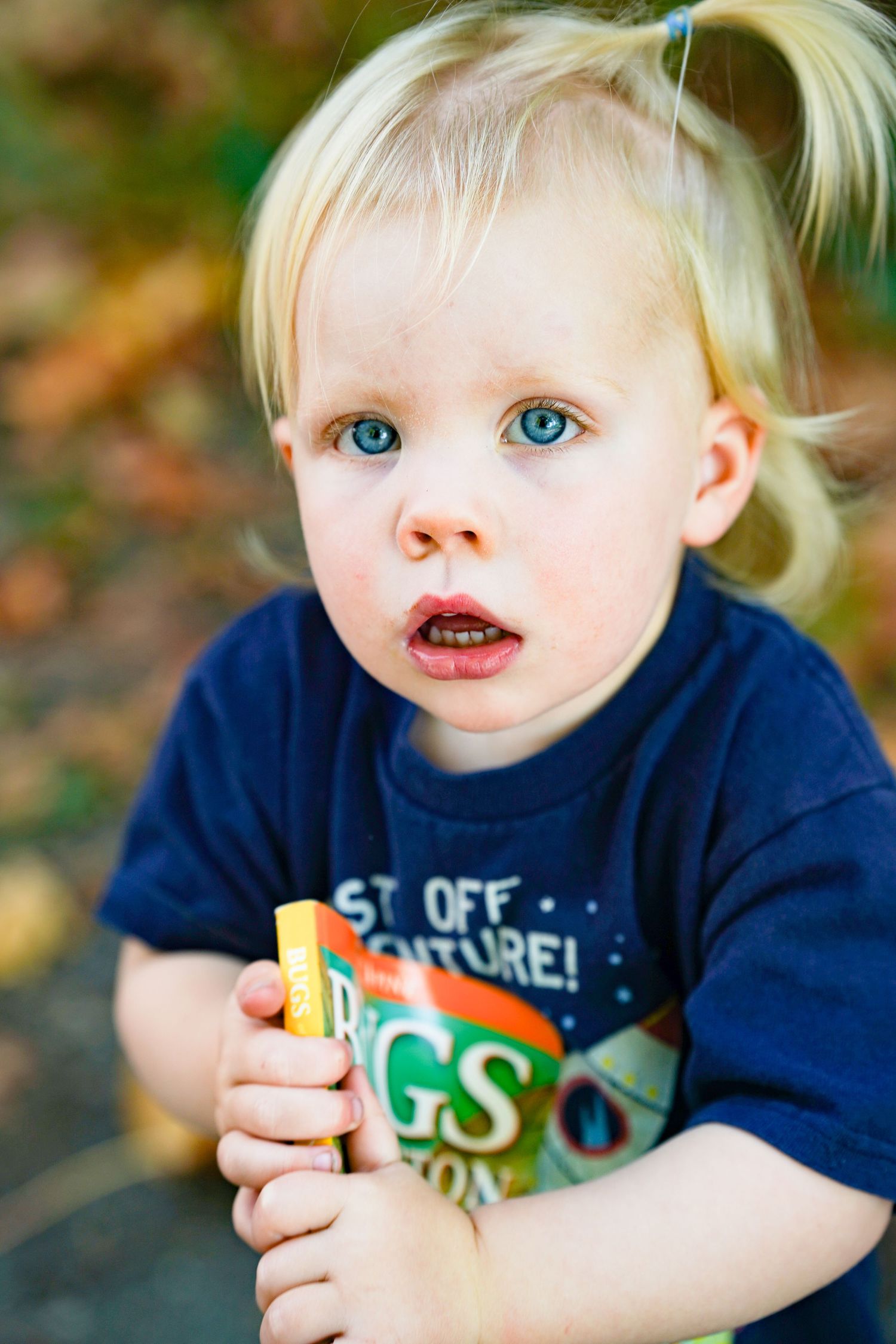 Blonde toddler with blue eyes holding a snack, wearing a navy blue t-shirt outdoors in autumn.