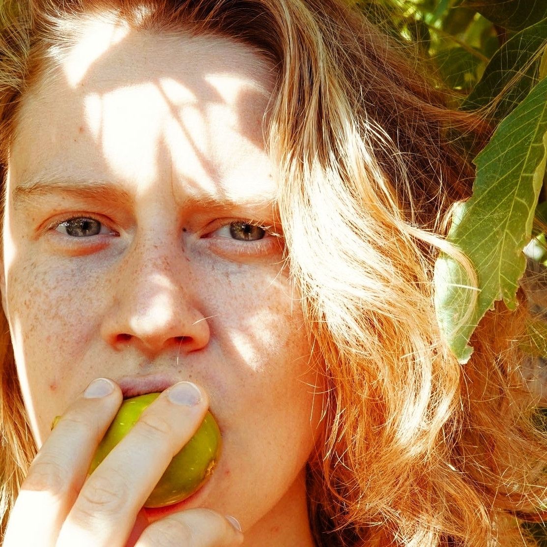 A photo of a woman with freckles in the sunlight and eating a green fig.