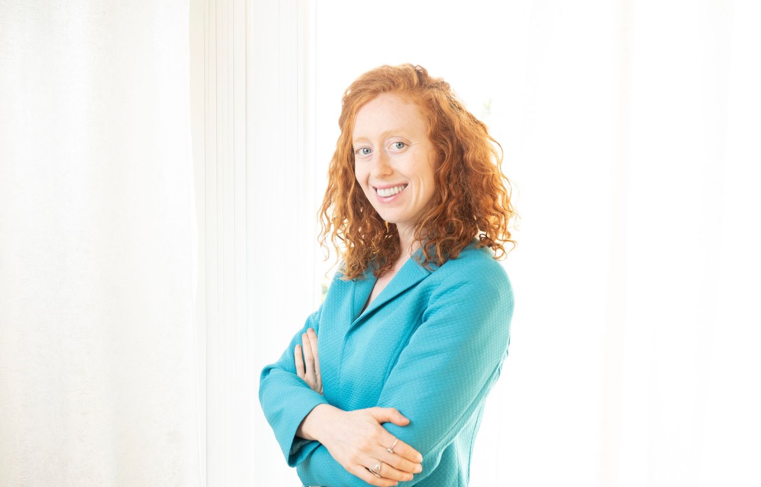 Smiling redhead woman in teal blazer posing with arms crossed against white background.