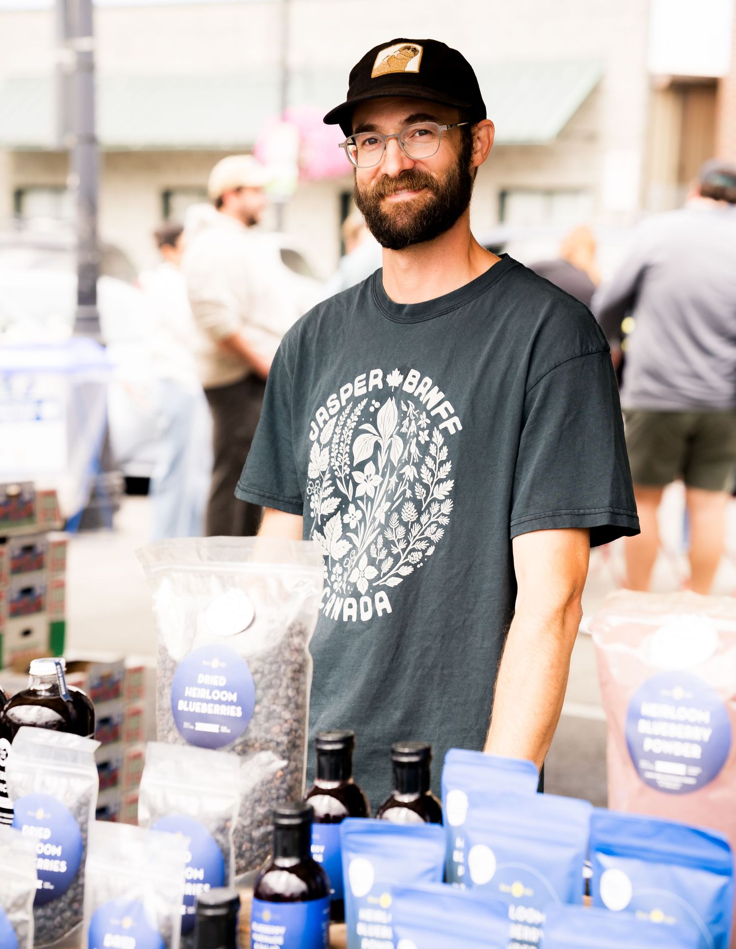 Bearded man wearing glasses and a dark graphic t-shirt stands at an outdoor market booth with bottles and packaged goods.