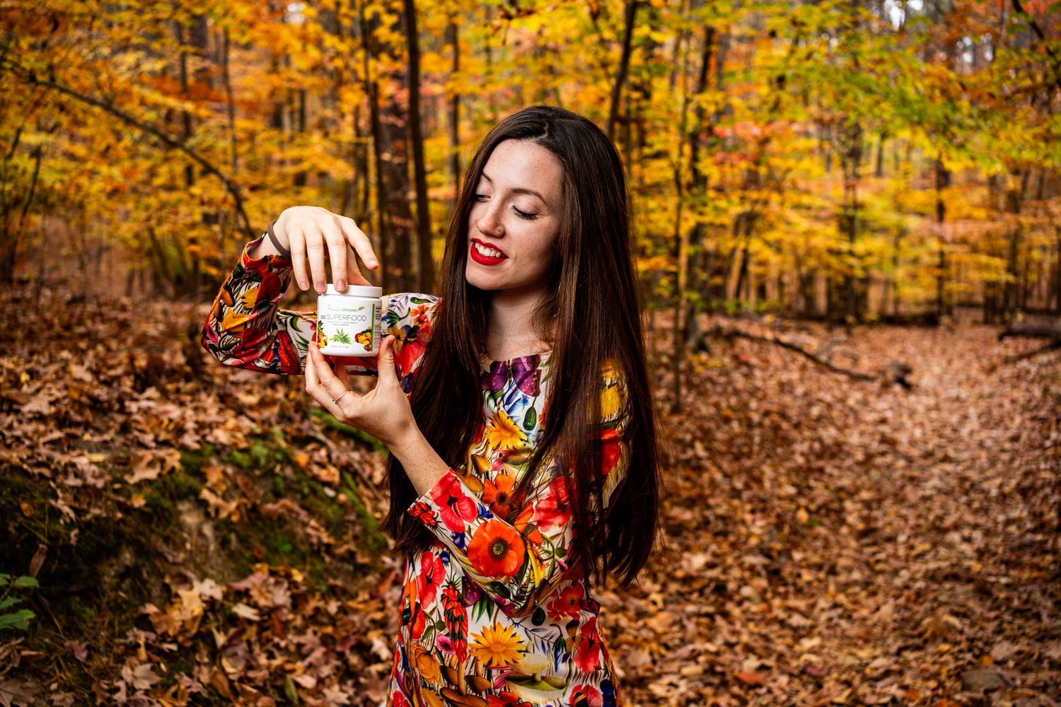Woman in colorful floral dress pouring tea in an autumn forest surrounded by golden fall foliage.