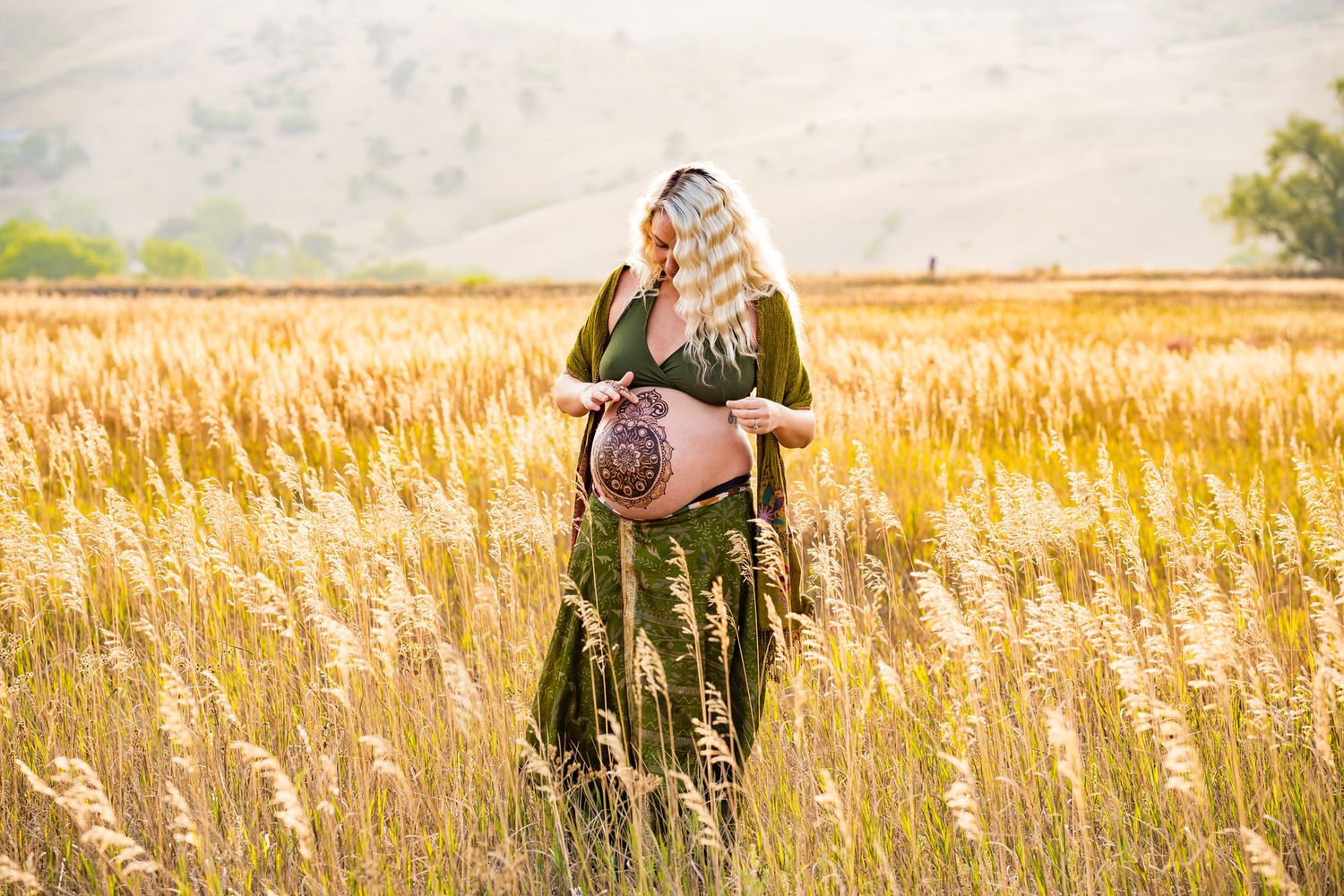 Pregnant woman with henna belly art standing in golden wheat field wearing green dress in warm sunlight.