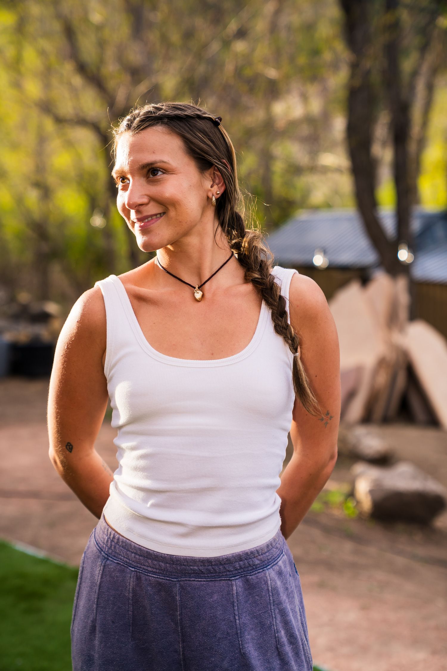 Smiling woman in white tank top and blue skirt standing outdoors in natural garden setting.