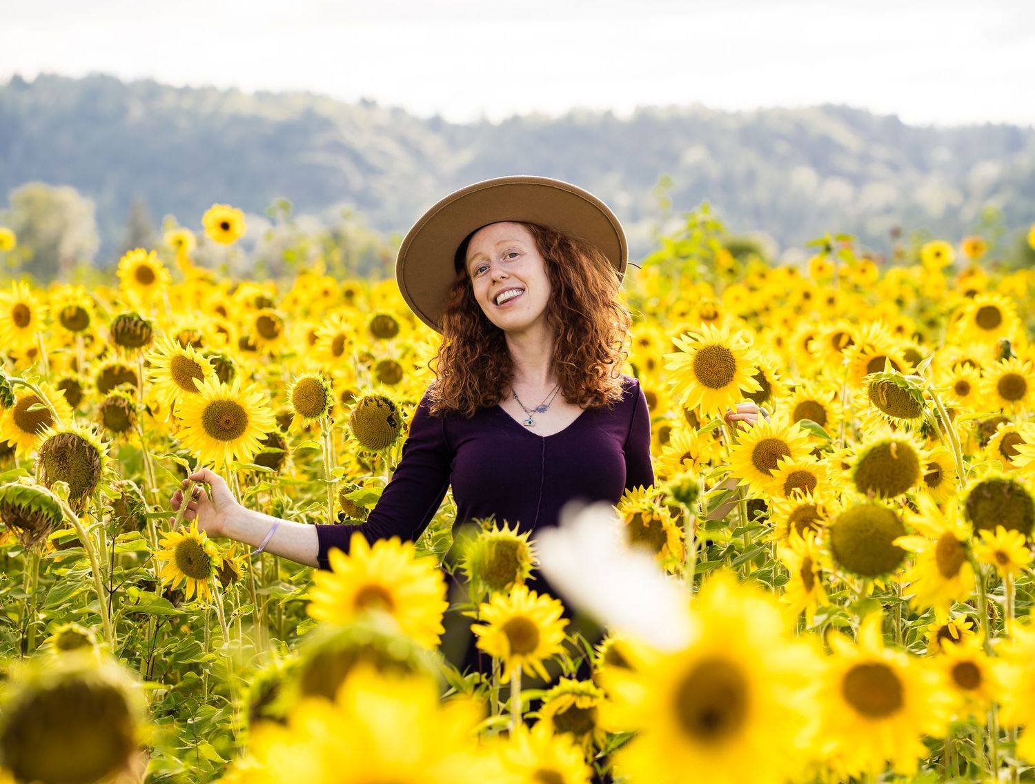 Woman in hat laughing joyfully in a vibrant sunflower field with lush green hills in the background.