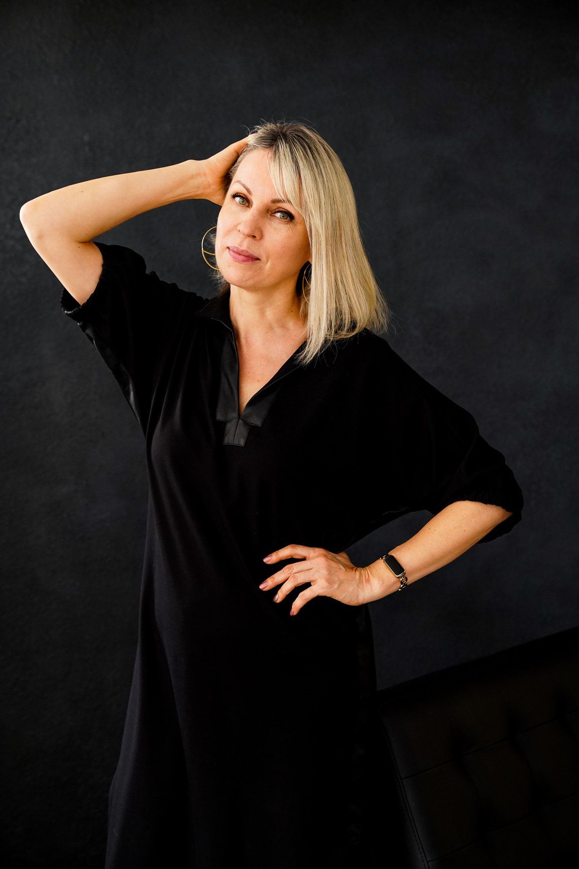 A photo of woman in the studio with a black dress and black background with hand on her head.