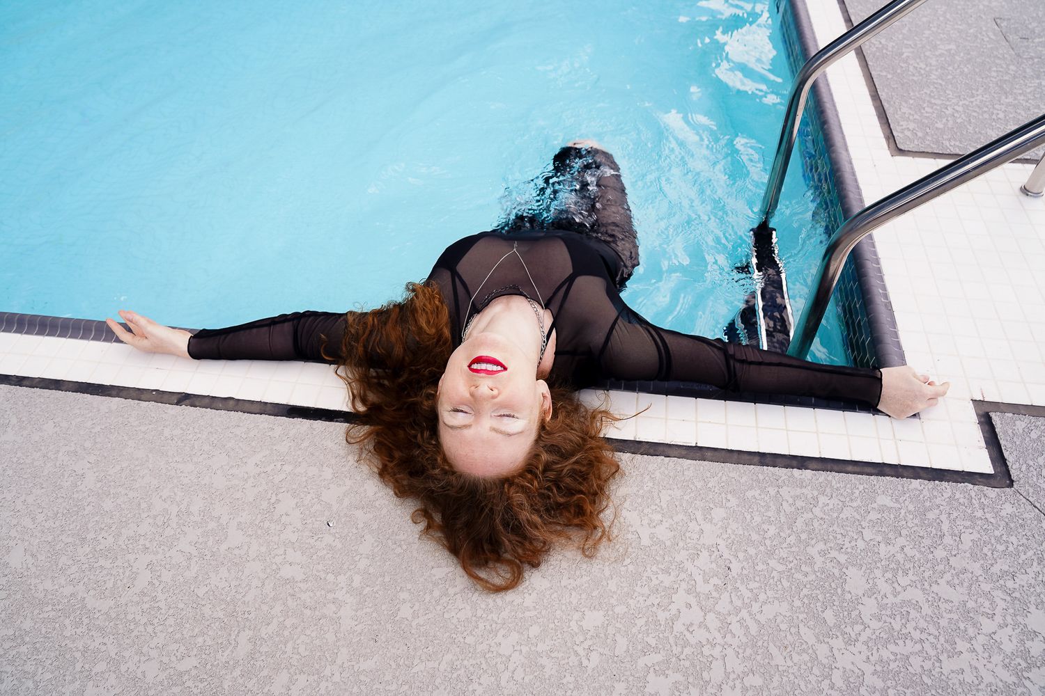 A photo of a girl with red hair and bright red lipstick laying in the pool with her arms out.