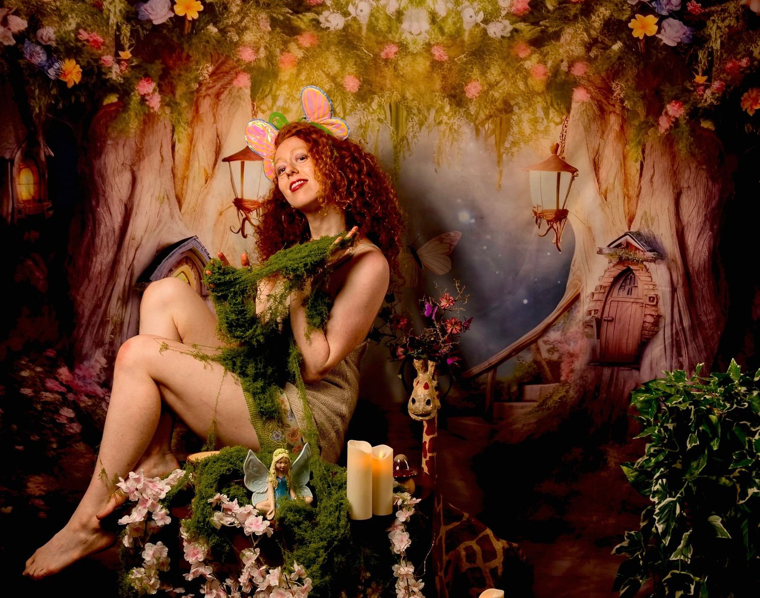 A woman with red curly hair poses as a fairy in an enchanted forest setting with candles, flowers, and a whimsical backdrop.