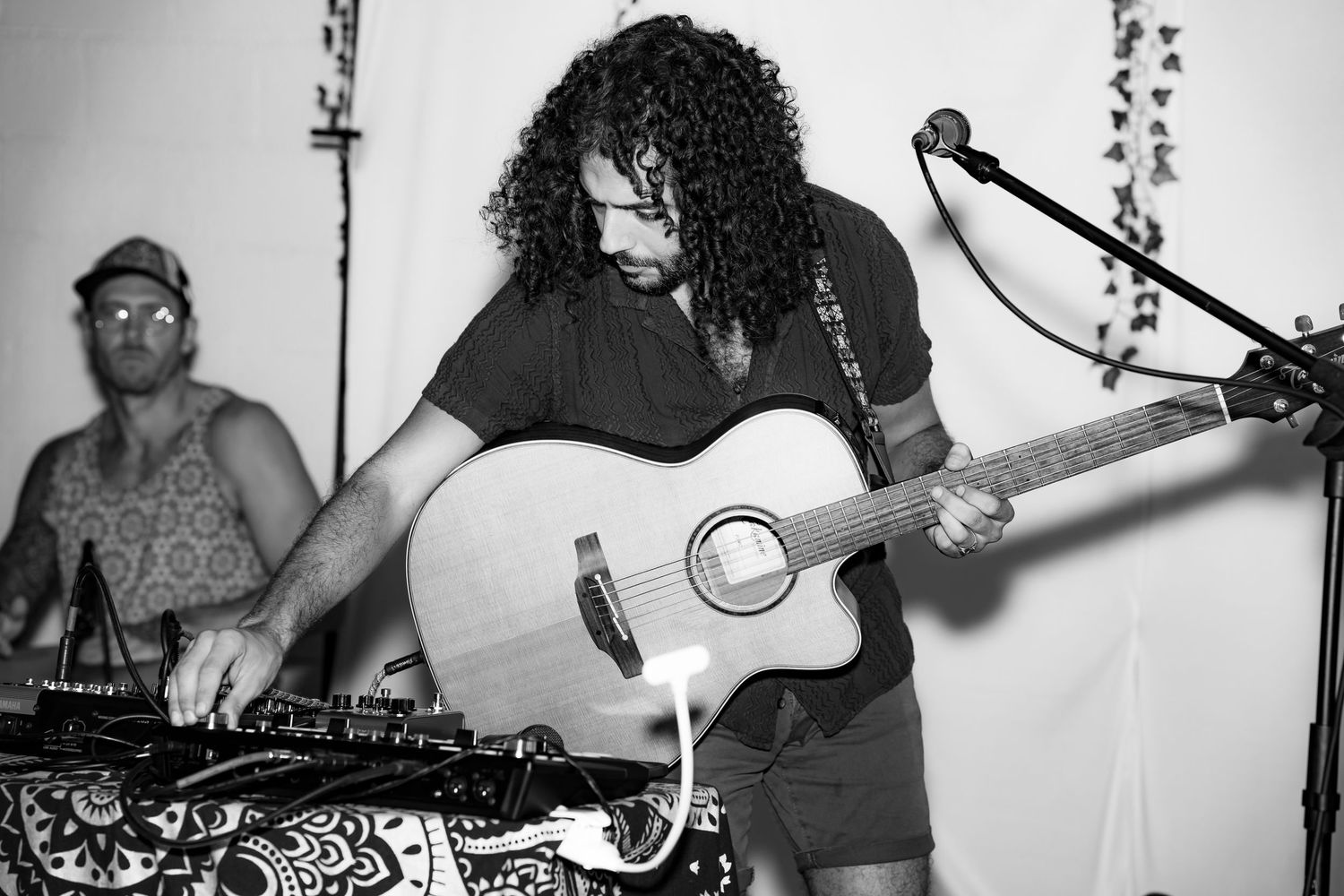 A photo of a man working on the sound board while holding his acoustic guitar.